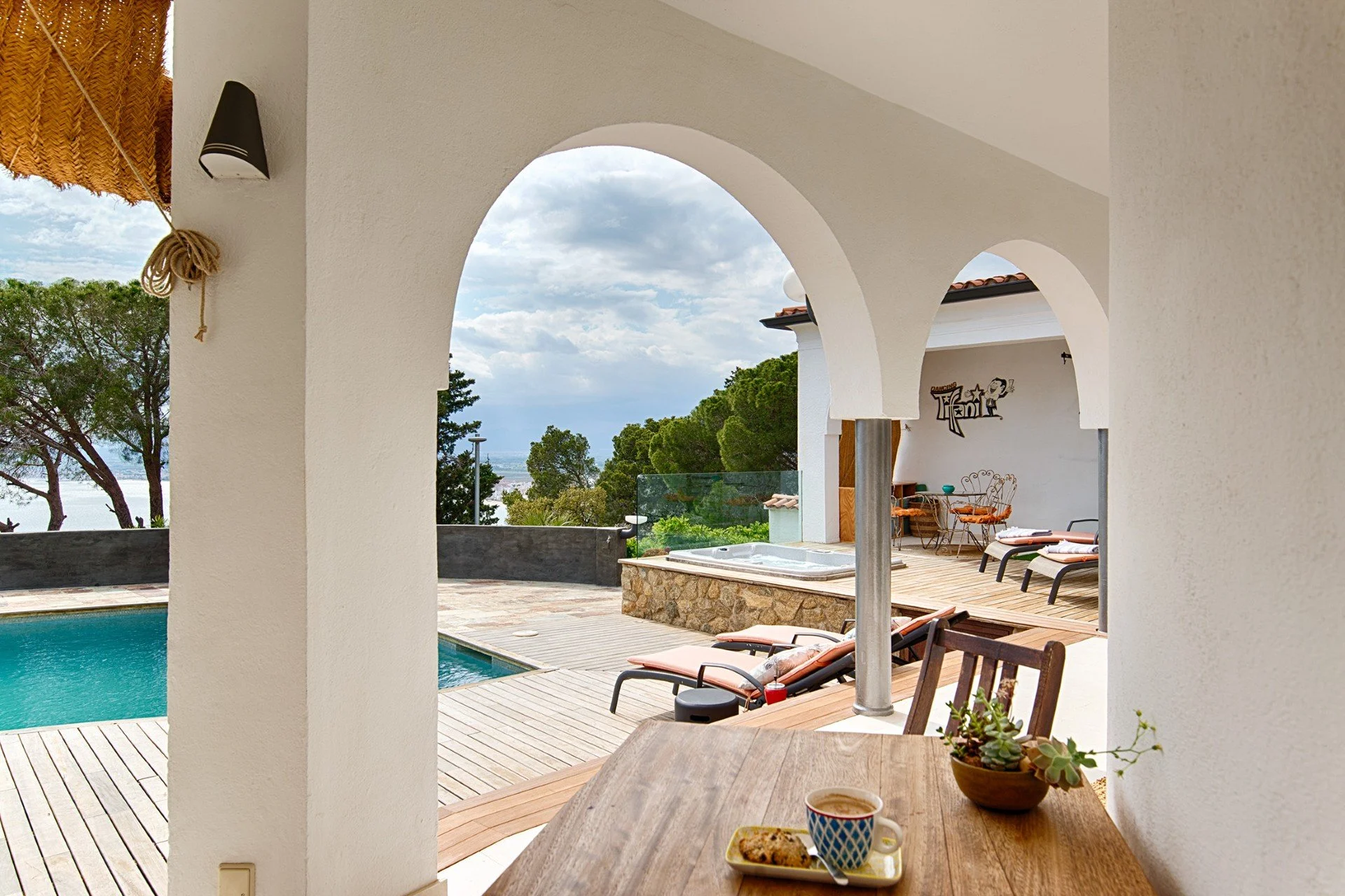 View of an outdoor patio with a swimming pool, lounge chairs, and a hot tub, framed by white arches, overlooking trees and a cloudy sky.