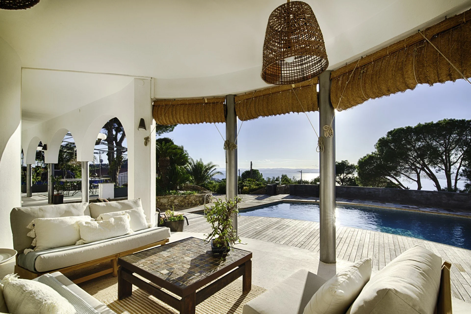 Outdoor living area with white cushioned seating, a wooden table, potted plants, and a view of a swimming pool and trees in the background, under a shaded canopy with woven hanging lamp.