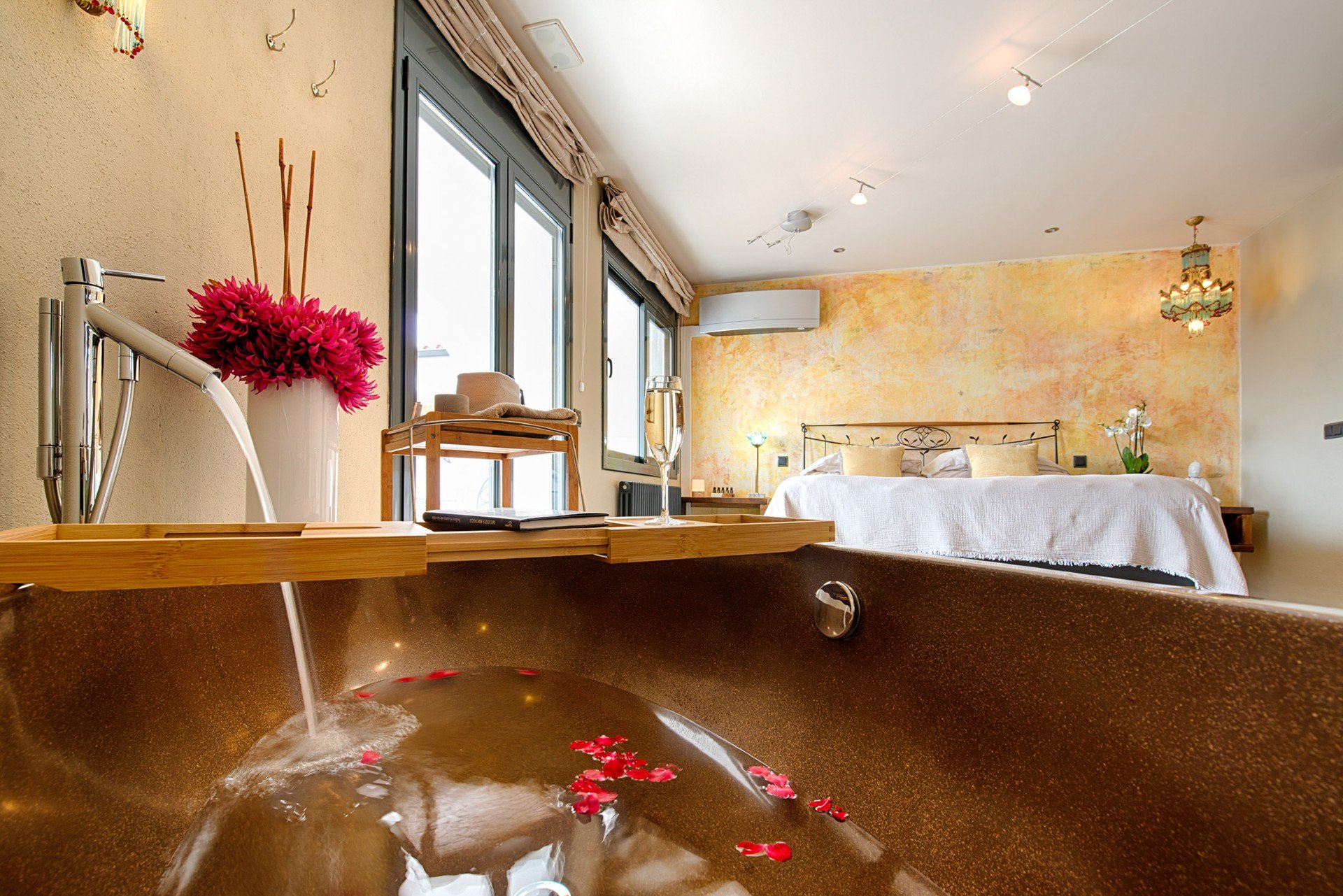 The King Room at Casa de l'Albada, Spain. A hotel room with a bathtub filled with water and red flower petals, a vase with pink flowers, a glass of champagne, a bed with gold pillows, large windows with curtains, and a decorative chandelier.