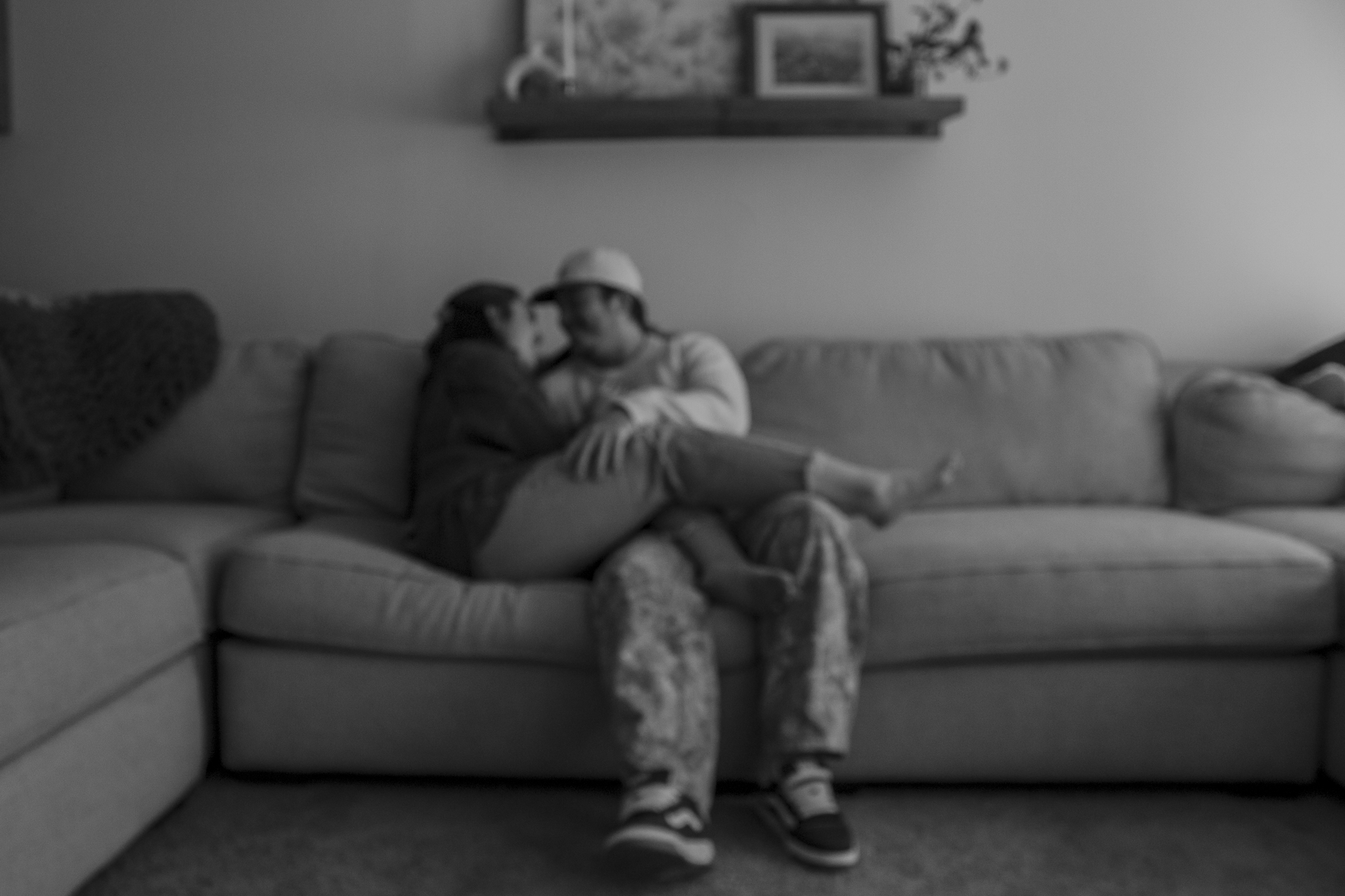 A person sitting on a couch, wearing a safety helmet, with their face close to a child. The person's face is blurred, and the child wears a cap. The scene is in a living room with framed pictures on the wall and a shelf. The person's legs are crossed, and they are wearing sneakers and casual clothing.