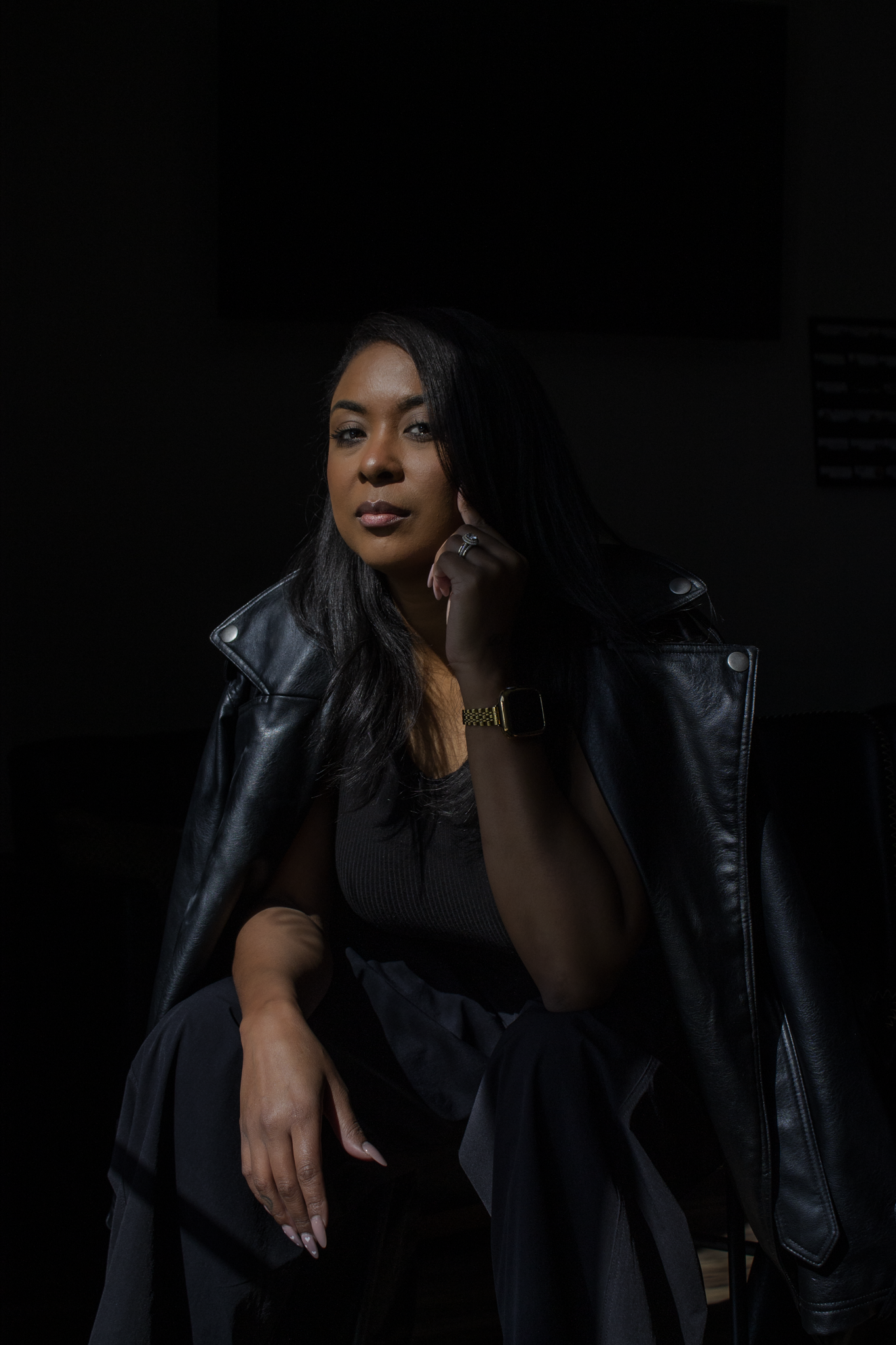A woman with long dark hair wearing a black leather jacket and black outfit, sitting against a dark background, with one hand resting on her chin and the other on her knee, looking into the camera with a neutral expression. Real estate firm branding.