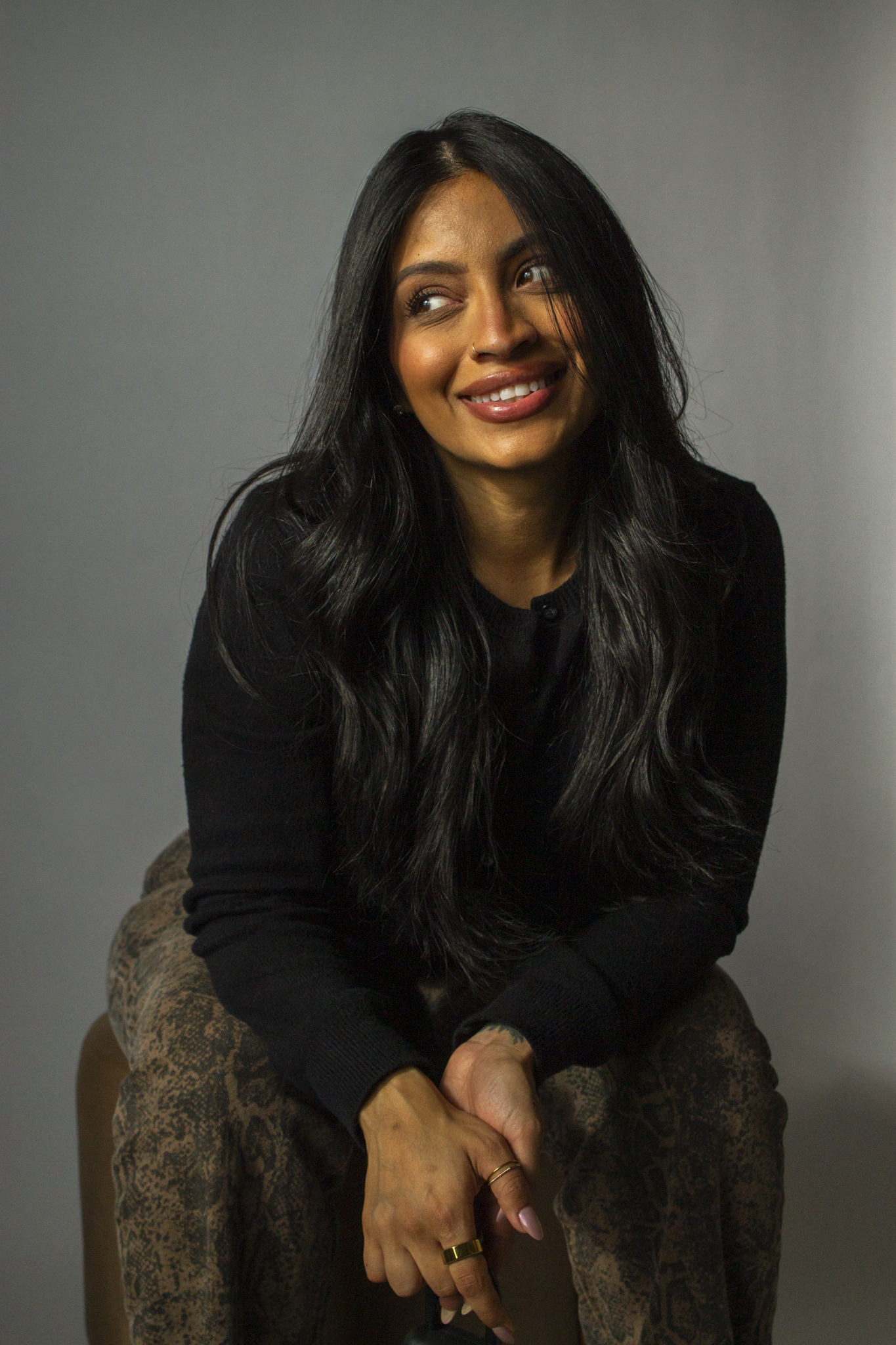 A woman with long black hair, wearing a black sweater and patterned pants, sitting on a chair against a gray background. posed darker branding shoot for real estate agent and business owner