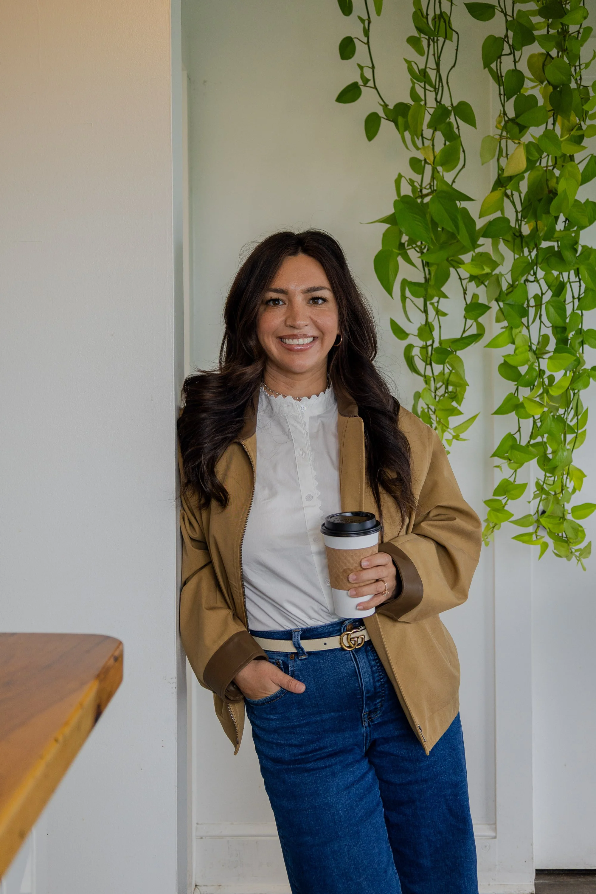 A woman with long dark hair holding a coffee cup, smiling, standing in front of a green leafy plant and a white wall in an indoor setting. branding shoot for realtors.