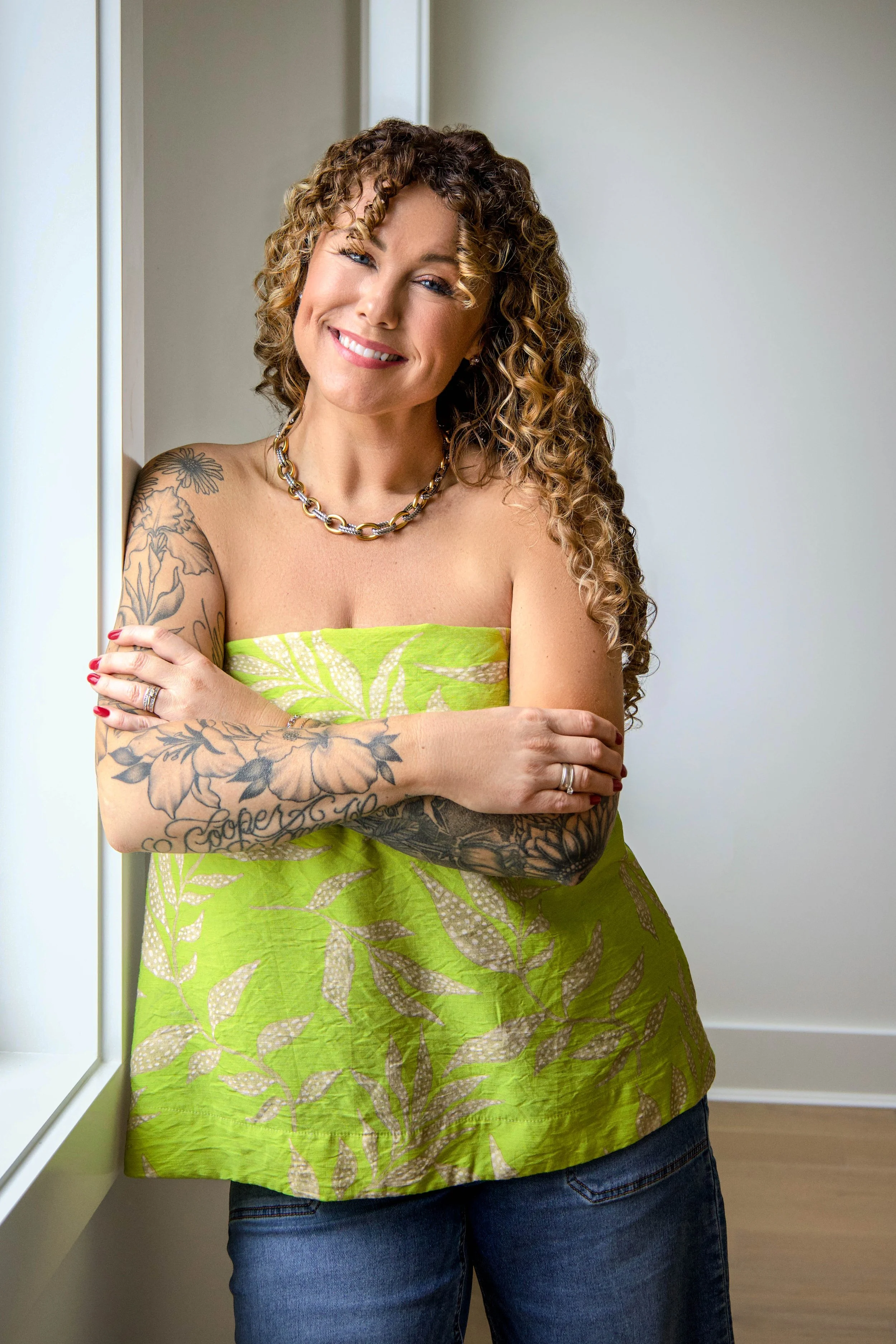 A woman with curly hair and tattoos on her arms, wearing a neon green strapless top with a leaf pattern, standing near a window with her arms crossed, smiling at the camera. new construction branding shoot photography. neutral background house.