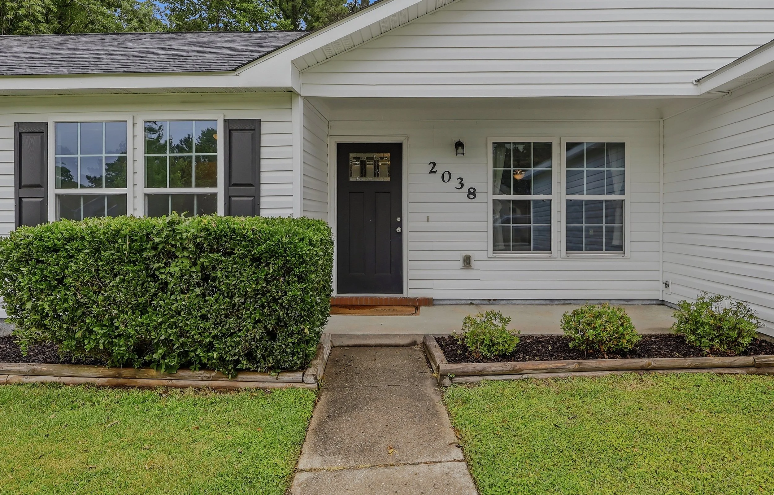 Front view of a single-story house with white siding, black shutters, and a black front door with house number 2038. There is a small concrete walkway leading to the door, surrounded by small bushes and a lawn.