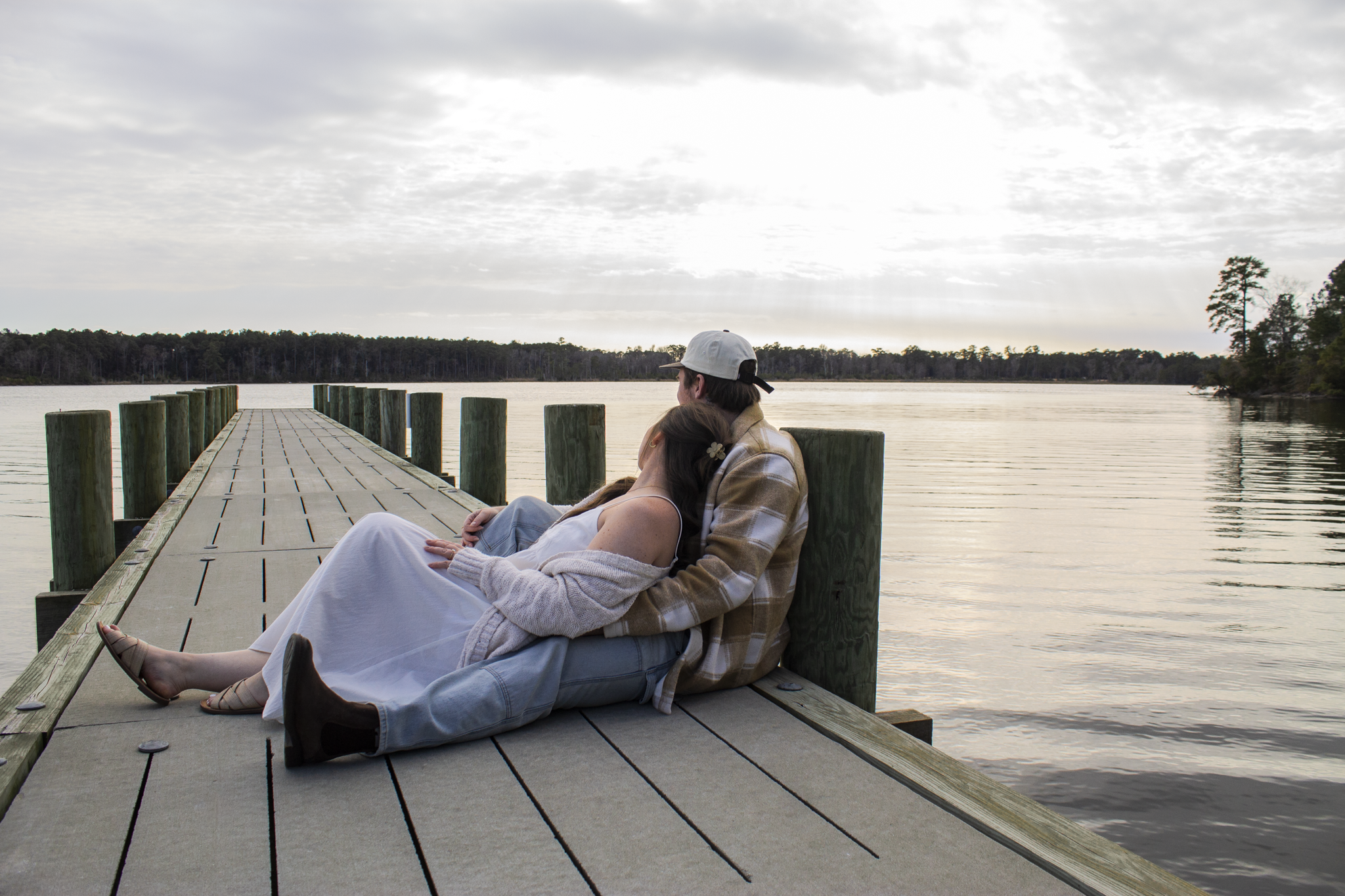 A couple sitting on a dock by a lake, embracing each other while facing the water under a cloudy sky.