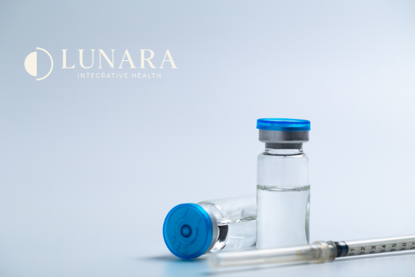 Two small glass vials of peptides with blue caps and a syringe lying in front of them, on a white surface with a logo for Lunara Integrative Health in the upper left corner.