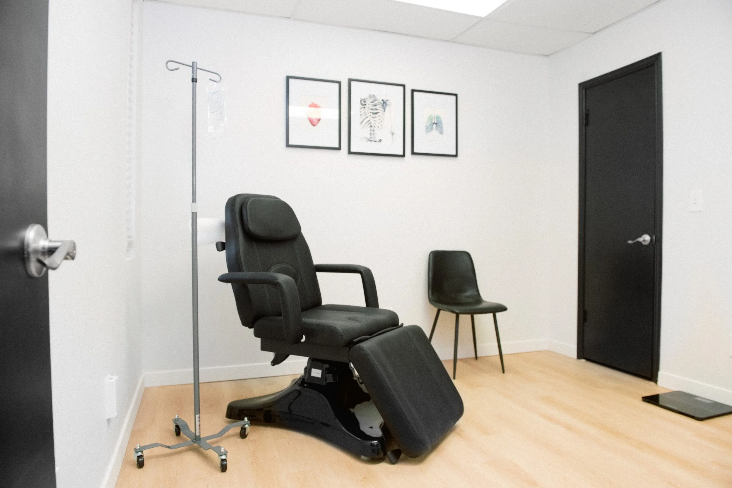 A warm medical examination room with a black doctor's chair, an IV stand with a bag, a black chair, three framed pictures on the wall, a black door, and a scale on the floor.