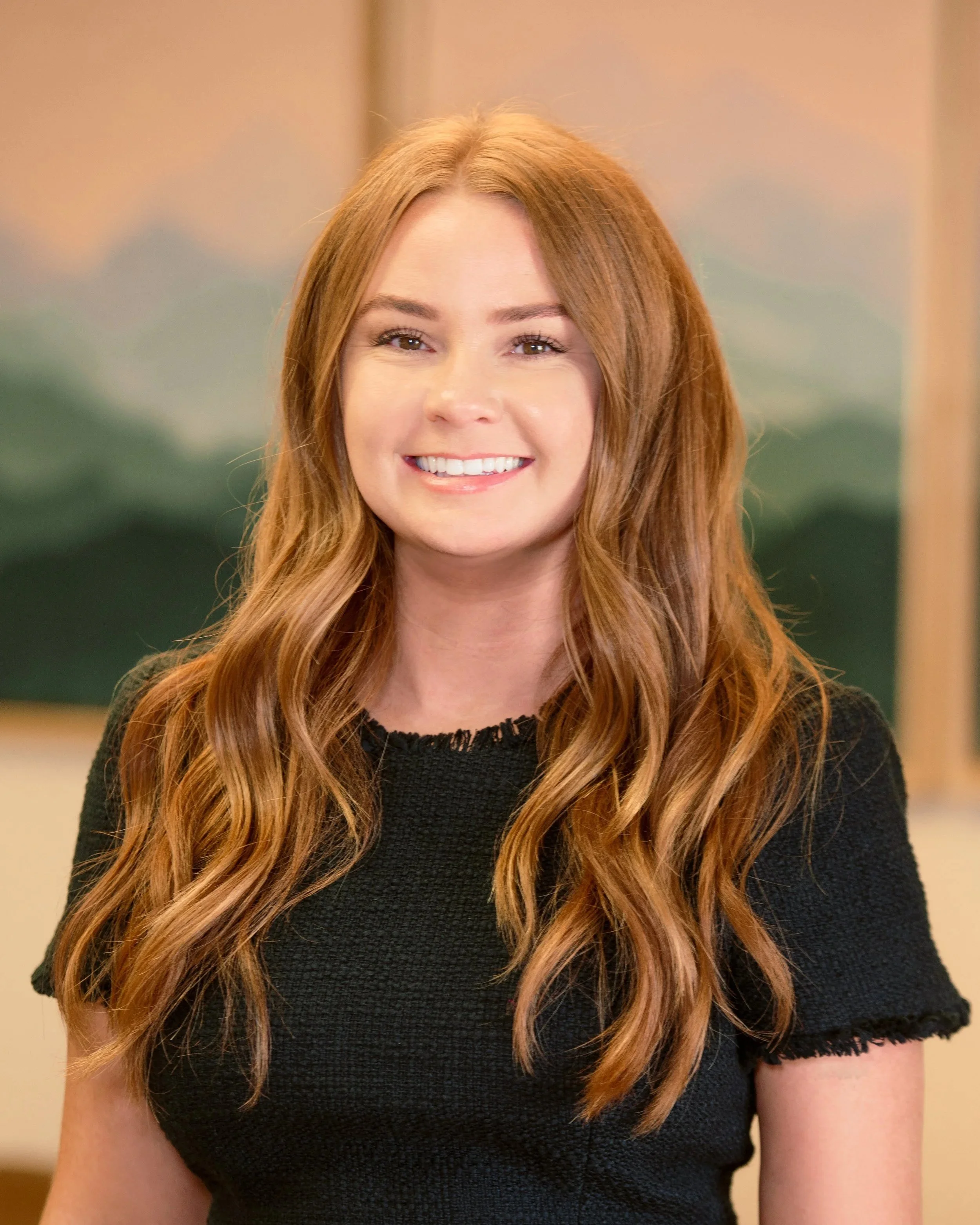 A young woman with long wavy red hair smiling at the camera, wearing a black short-sleeved top, with a blurred colorful background.