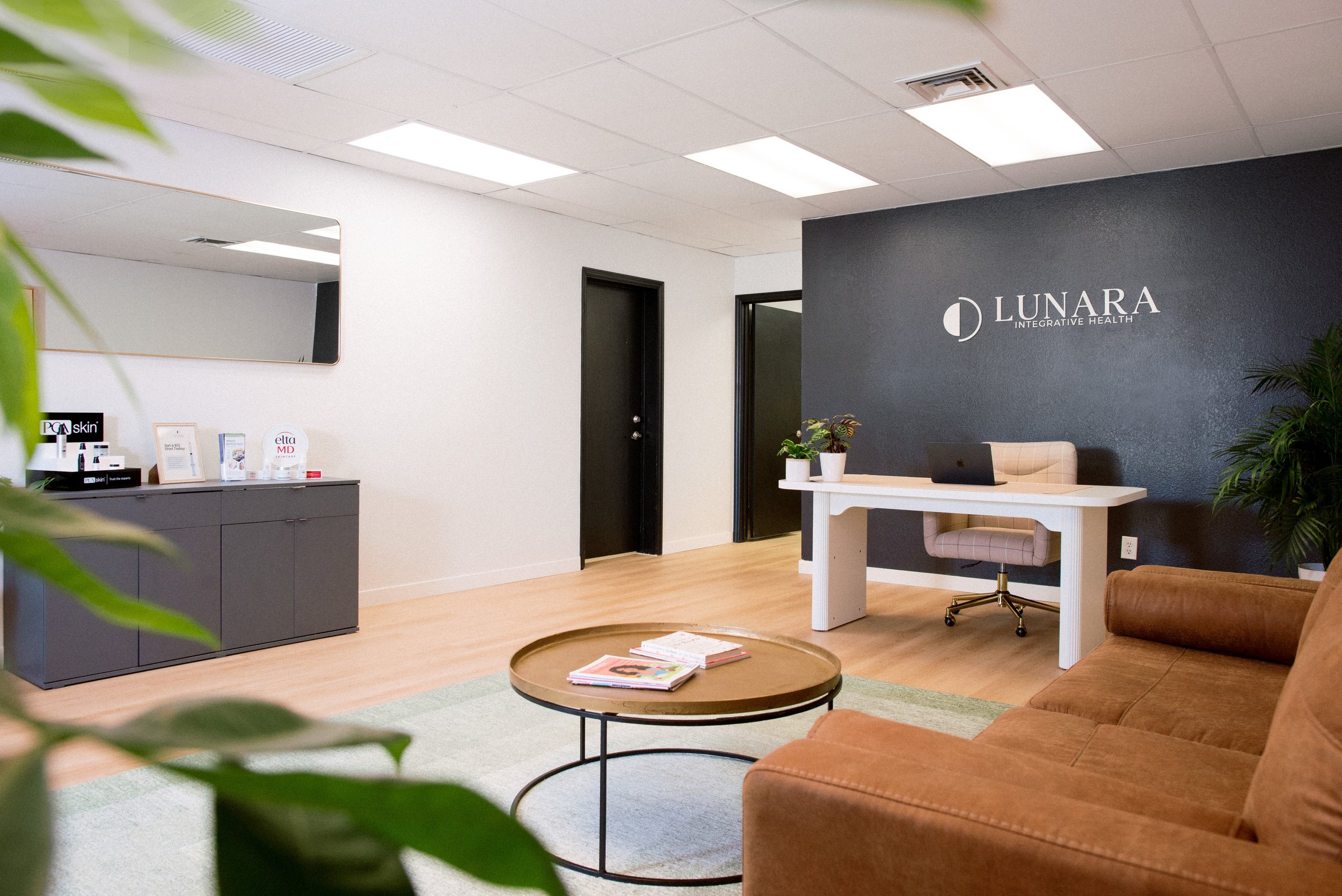 Comfortable reception area of Lunara Integrative Health with a brown couch, white desk with plants, a black wall with logo, and a mirror on white wall.