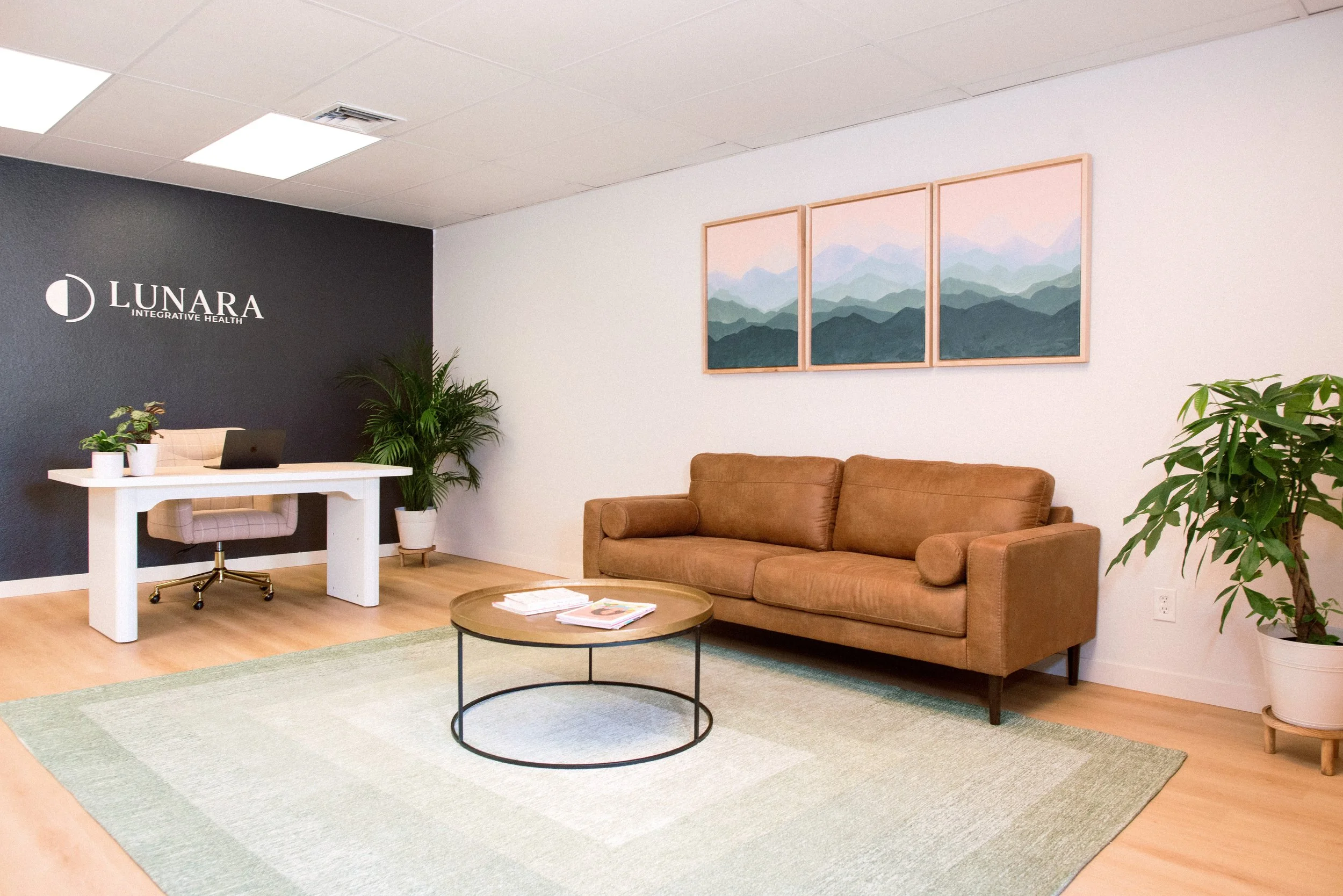 A peaceful office waiting room with a brown sofa, plants, and a white desk, featuring a logo 'LUNARA INTEGRATIVE HEALTH' on a dark accent wall.