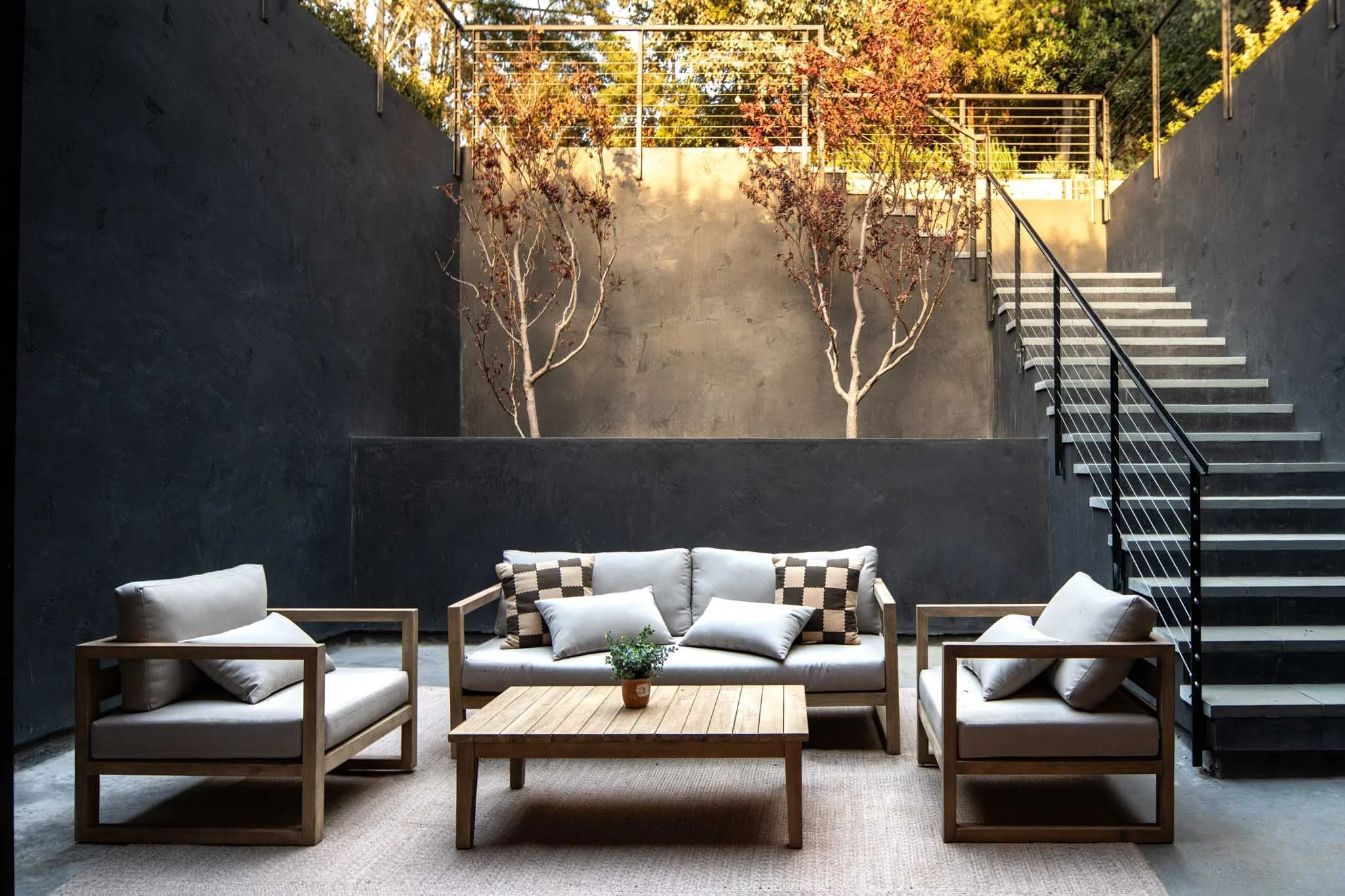 Modern outdoor patio with gray cushioned chairs and a matching sofa, a wooden coffee table with a small potted plant, black metal staircase, and two leafless trees against a black wall.