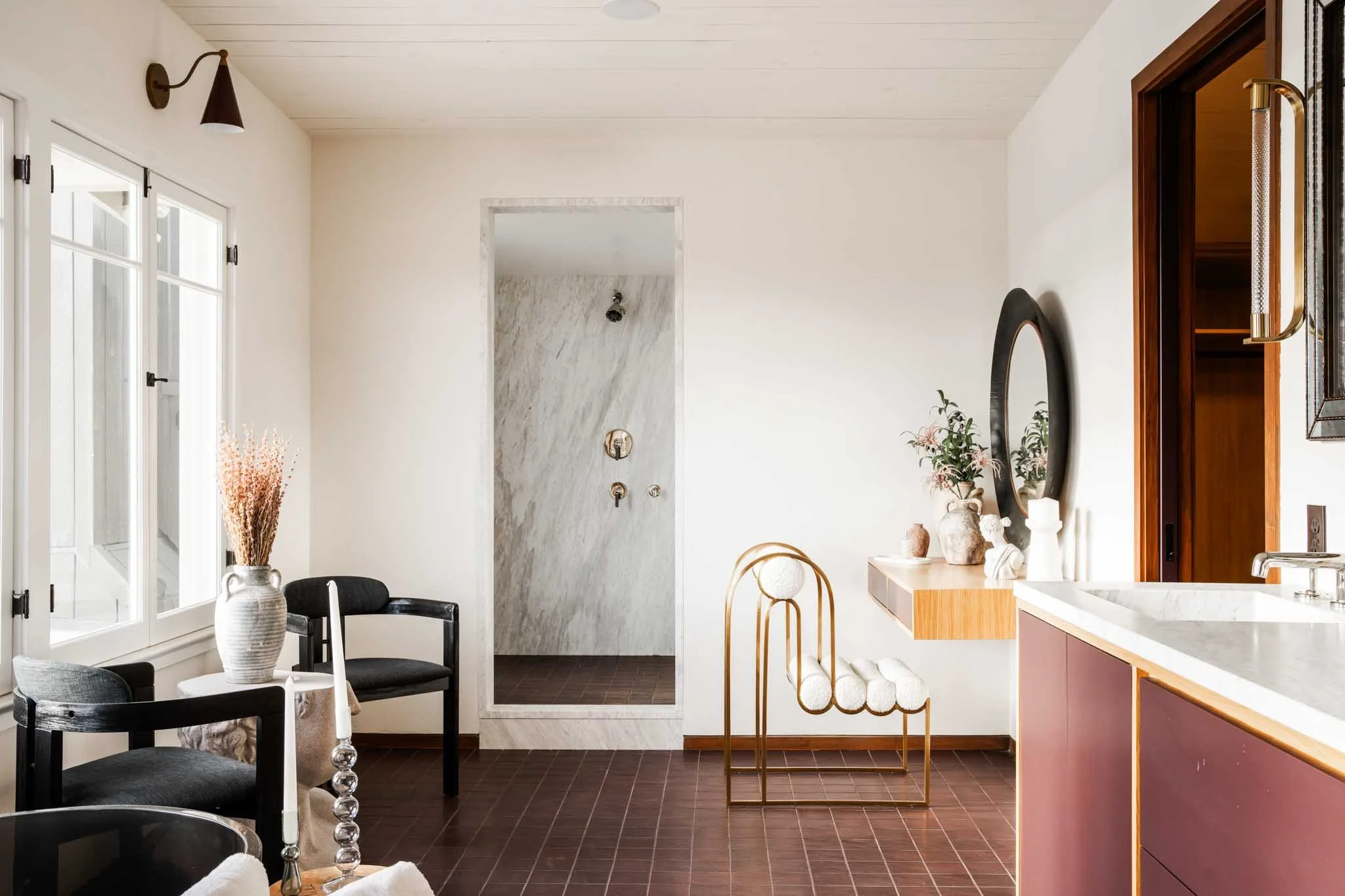 Bright, modern bathroom with white walls, dark wood flooring, a window with white framing, a black chair near a vase with dried pink flowers, a full-length mirror, a black-framed round mirror, a floating wooden vanity with pinkish-brown cabinetry, a marble countertop, decorative candles, pottery, and greenery, and a walk-in shower with marble tiles and brass fixtures.