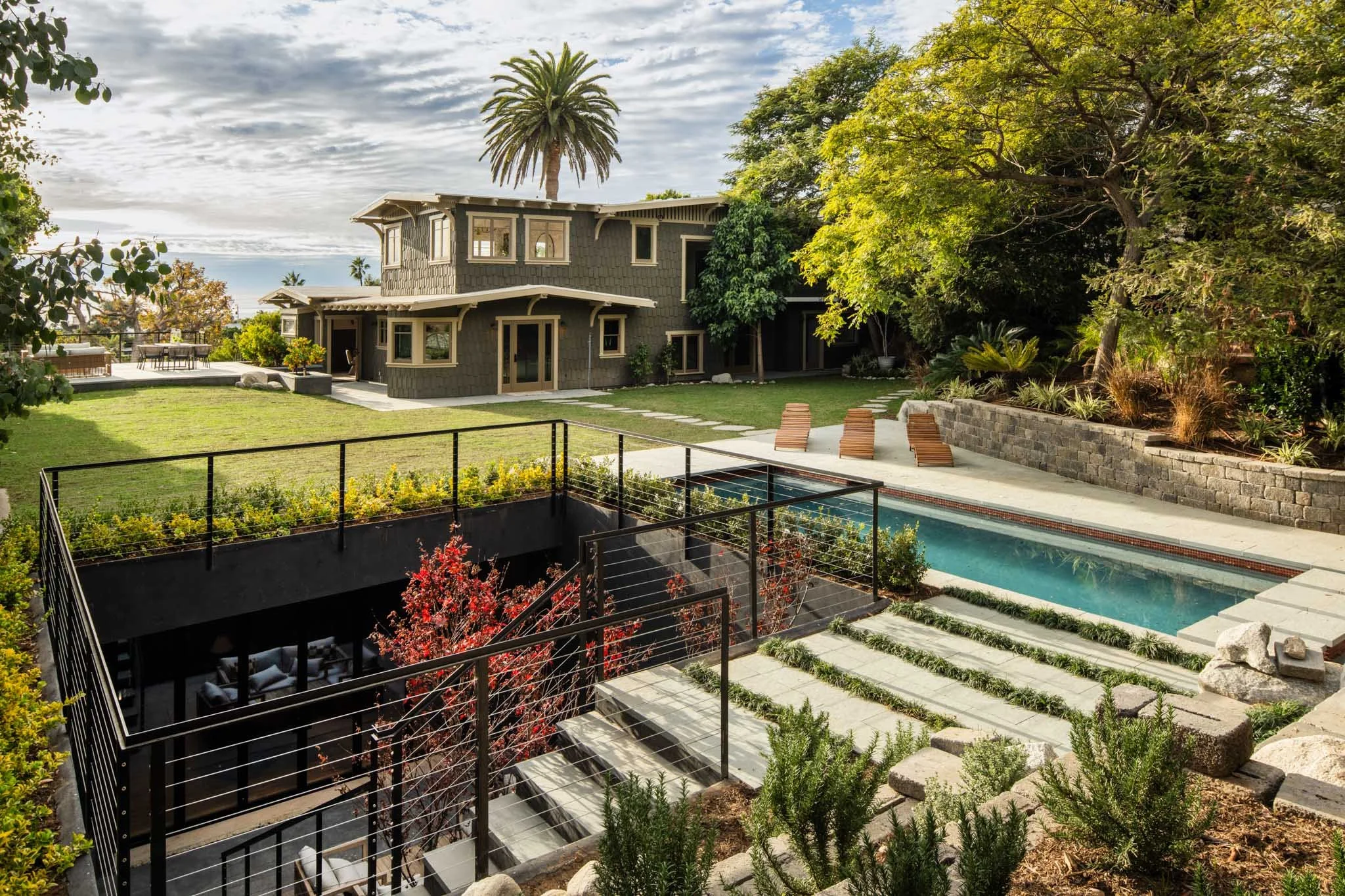 A modern backyard with a swimming pool, lounge chairs, a grassy lawn, and a two-story house with a deck and outdoor furniture surrounded by trees and landscaping.