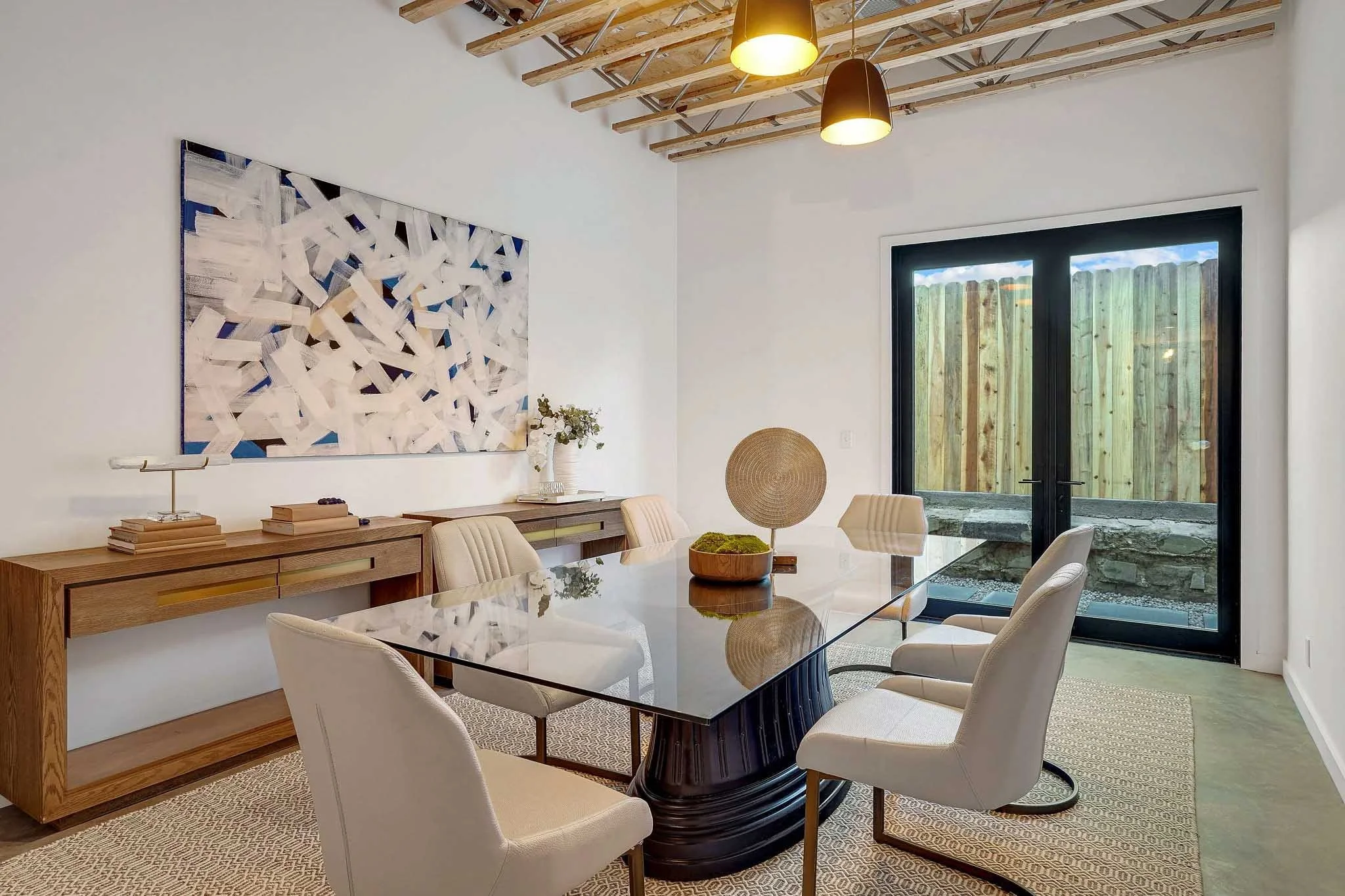 Modern dining room with glass-top table, six beige chairs, wooden sideboards, wall art, and large glass doors leading outside.