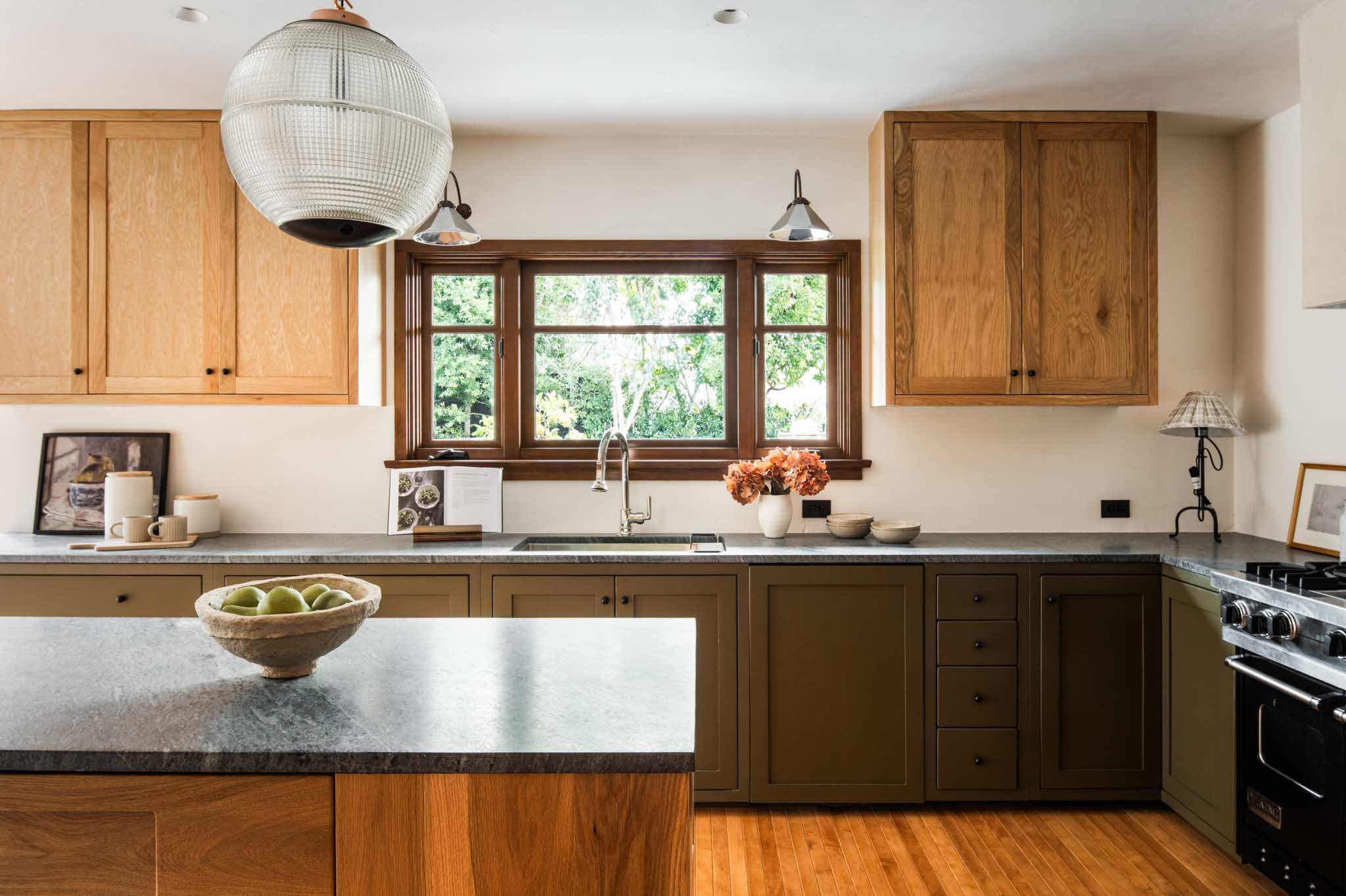 A dining room with a wooden table surrounded by eight beige upholstered chairs. A large, textured vase with white flowers is on the table. The room has wood paneling on the walls, two windows letting in natural light, and a modern chandelier hanging from the ceiling.
