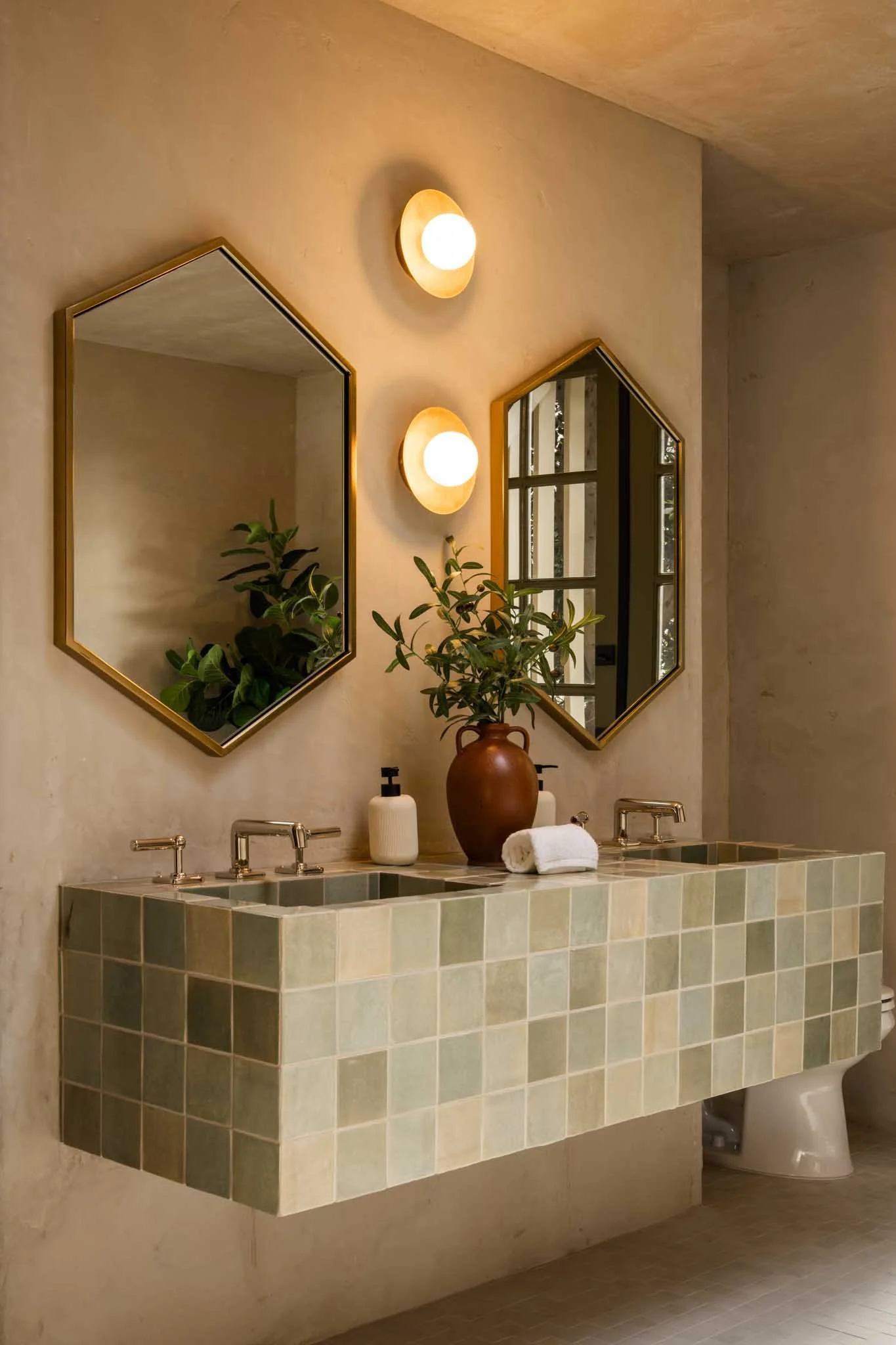 Bathroom with a green tiled double sink vanity, two mirrors, two wall-mounted lights, potted plant, soap dispensers, and a towel.