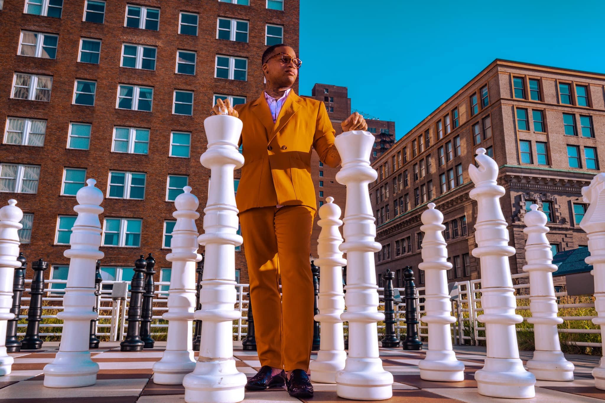 A man in a brown suit and sunglasses playing chess on an outdoor giant chessboard in an urban area with tall buildings in the background.