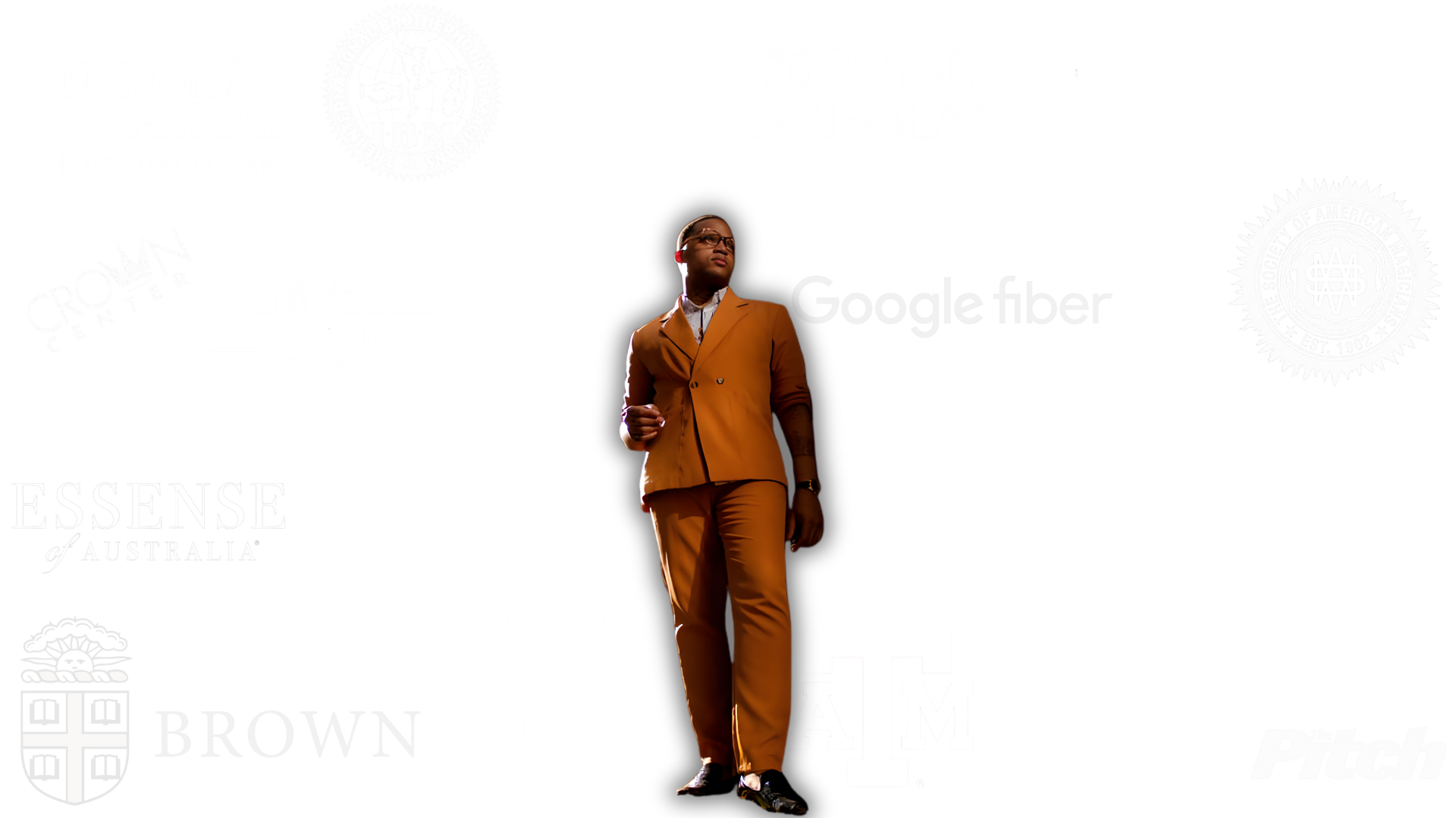 A person standing alone in front of a white background with various company logos and text, wearing a brown suit and black shoes, looking slightly to the side.