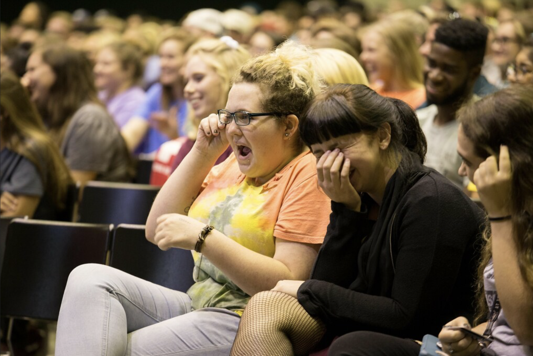 A group of people sitting in an audience, laughing and enjoying a performance or presentation. Two women in the foreground are laughing with their hands near their faces.