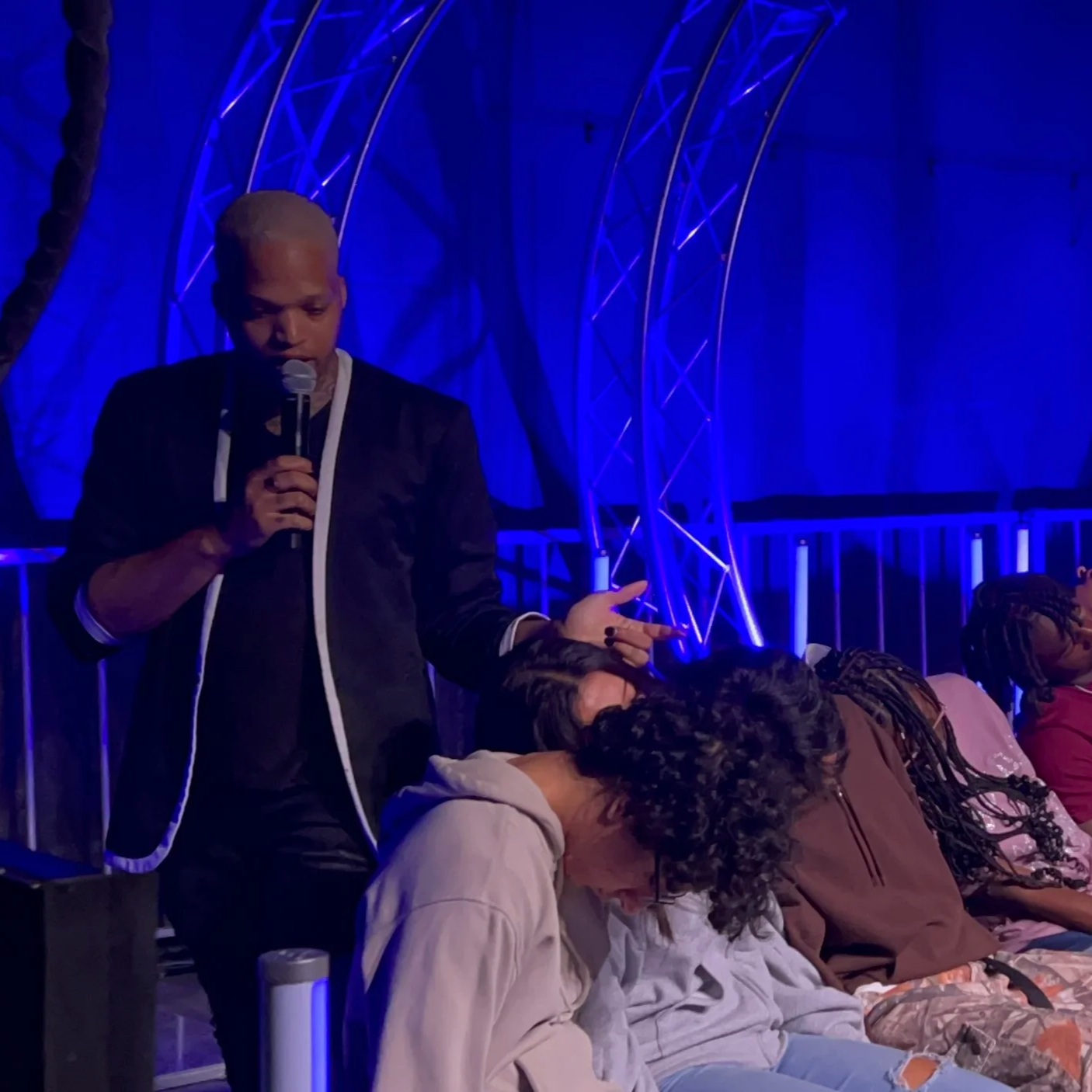 A person with short blond hair holding a microphone and speaking to a seated group in a dimly lit venue with blue lighting and metallic arch structures.