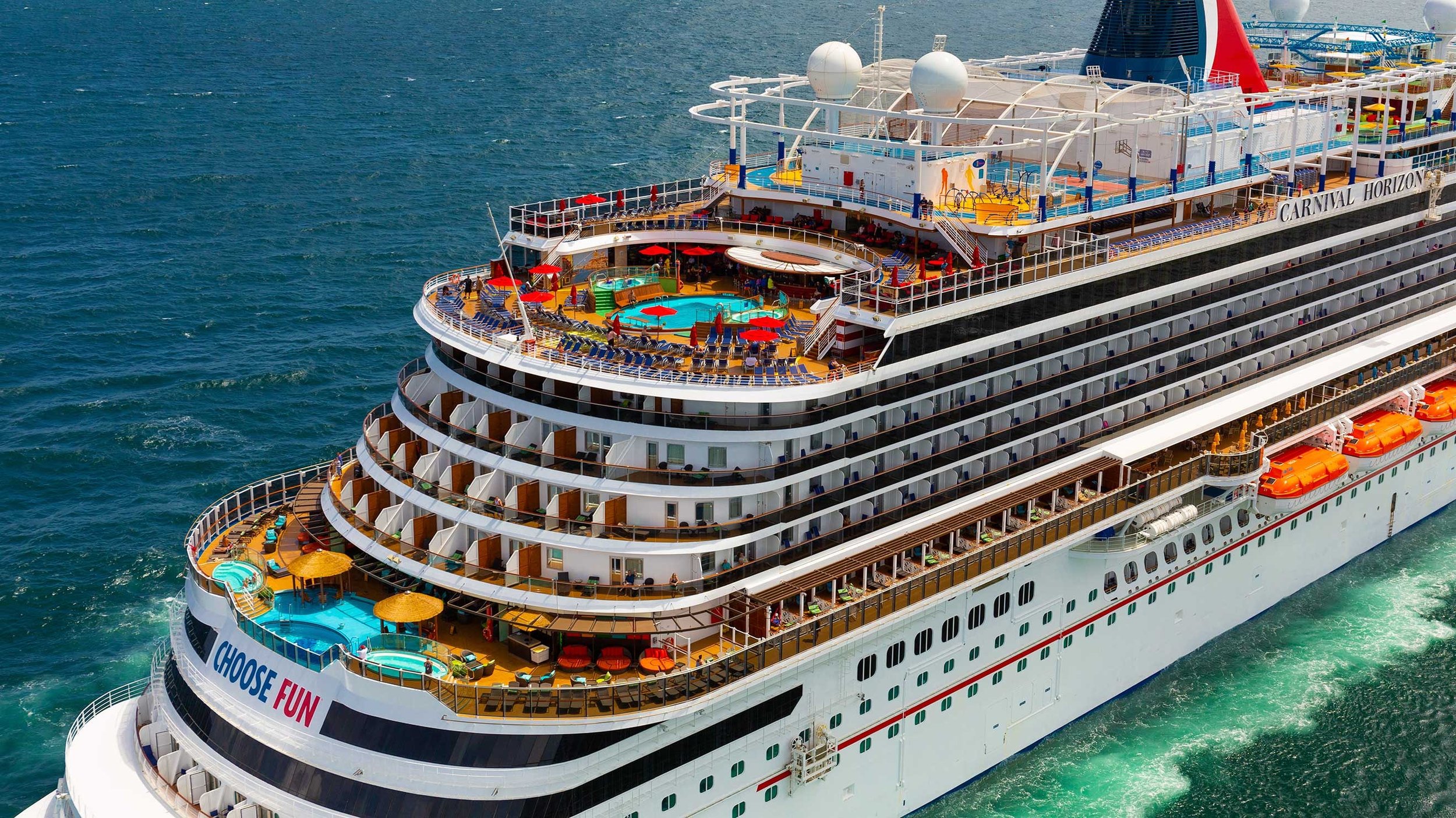 A large cruise ship on the ocean with multiple decks, pools, and outdoor amenities, including a hot tub, sun loungers, and shaded areas.