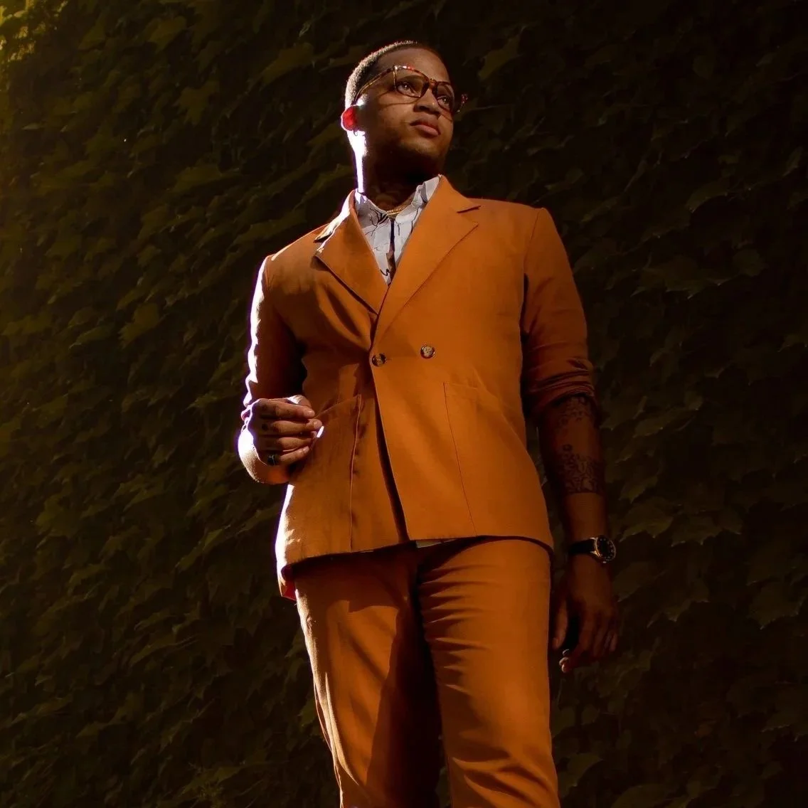 A man wearing a brown suit, glasses, and jewelry walking outdoors at night with a dark leafy background.