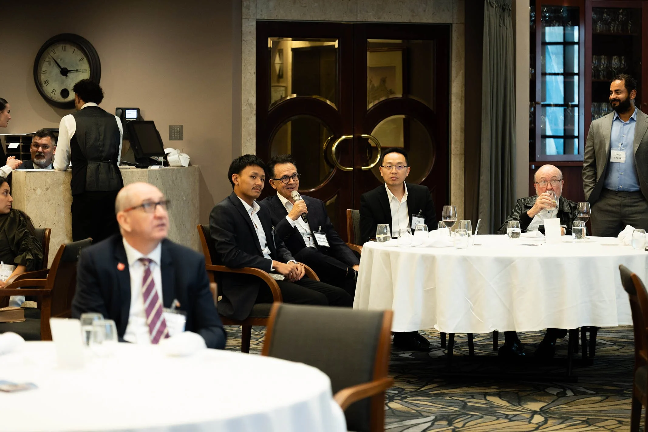Group of professionals attending a conference or meeting, seated at round tables with white tablecloths, in a formal setting.
