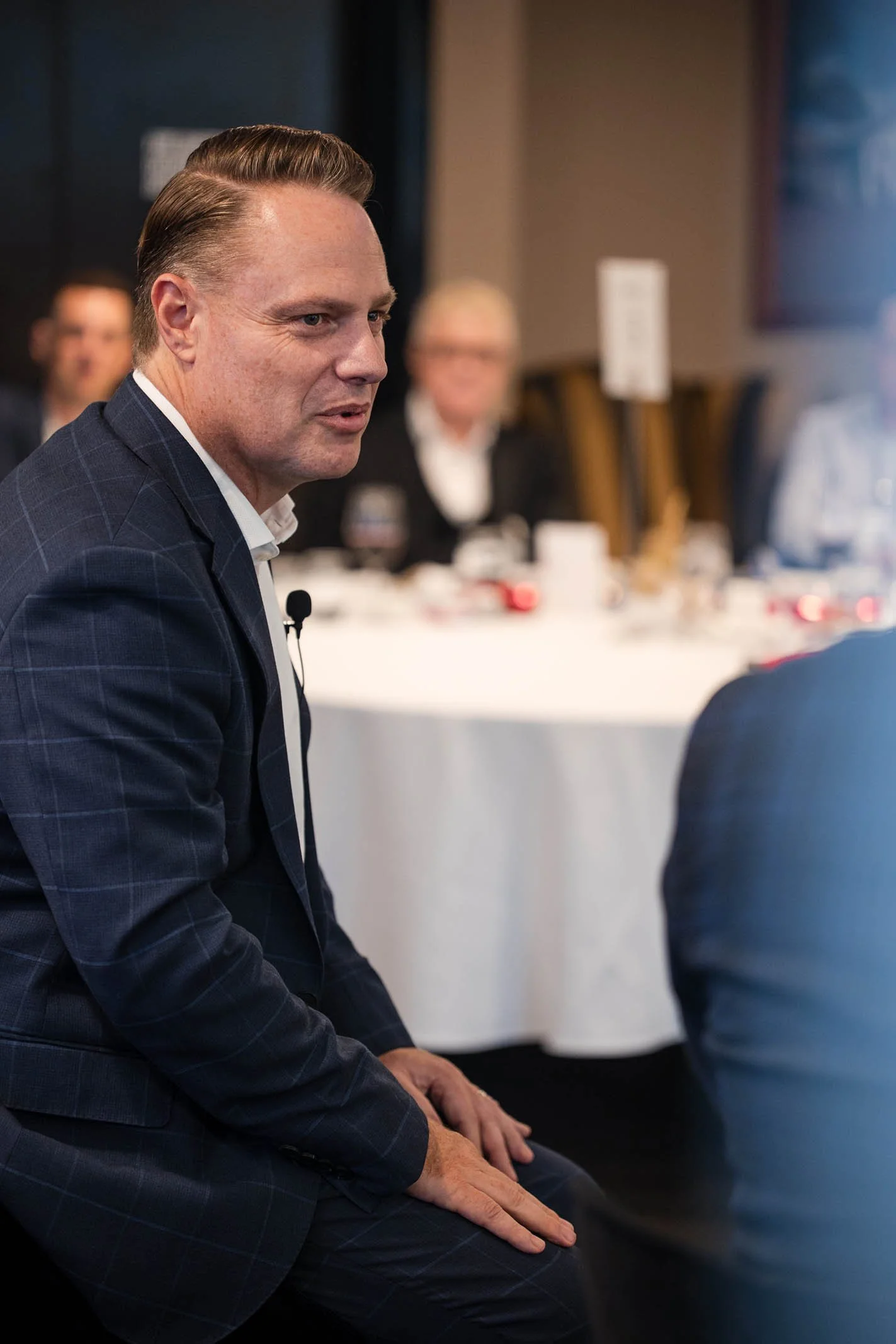 A man in a navy checkered suit engaged in conversation at a formal event, with a blurred background of other attendees and a round table with white tablecloths.