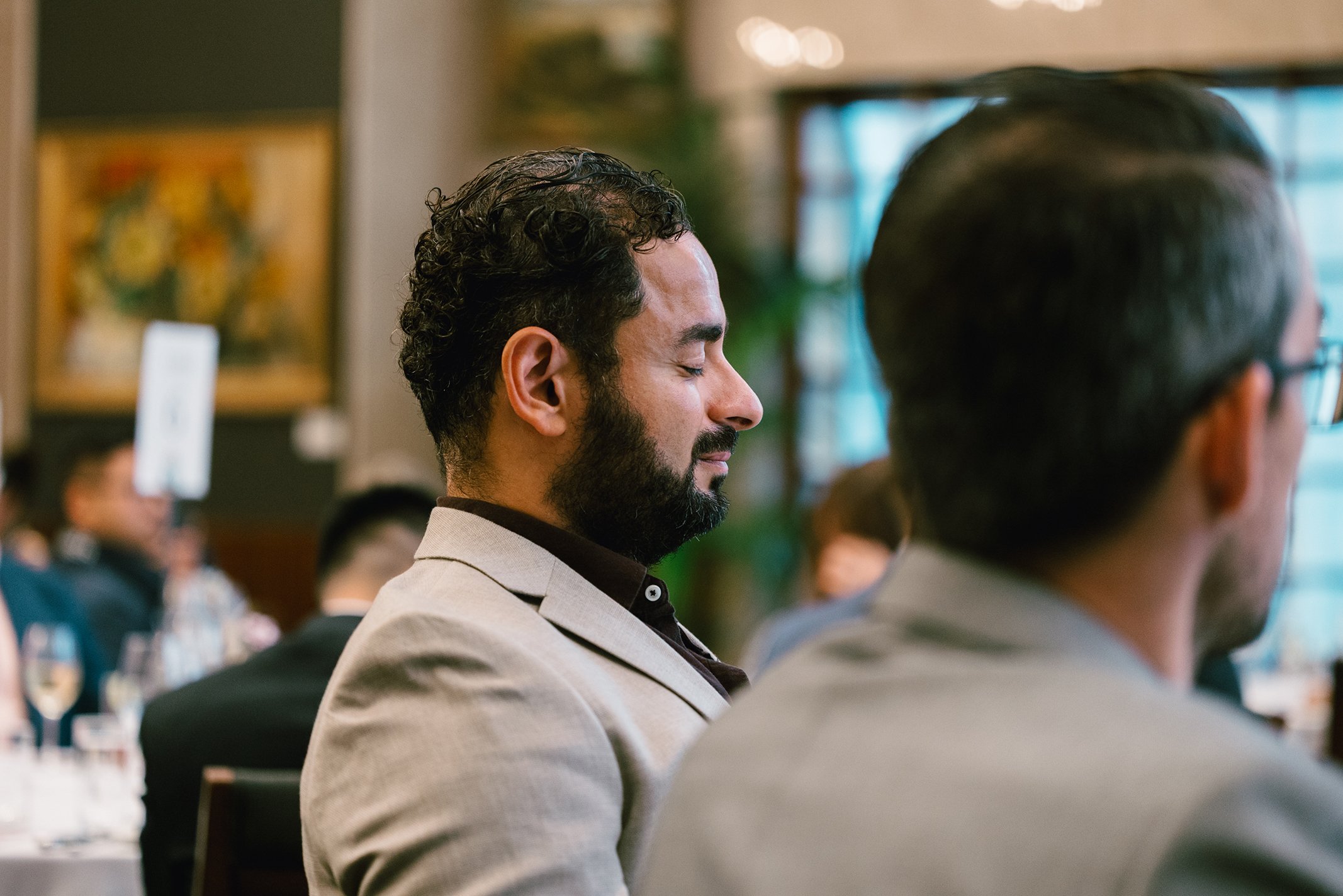 Man with curly hair and beard in a light suit, eyes closed, appearing to meditate or relax at a formal event.