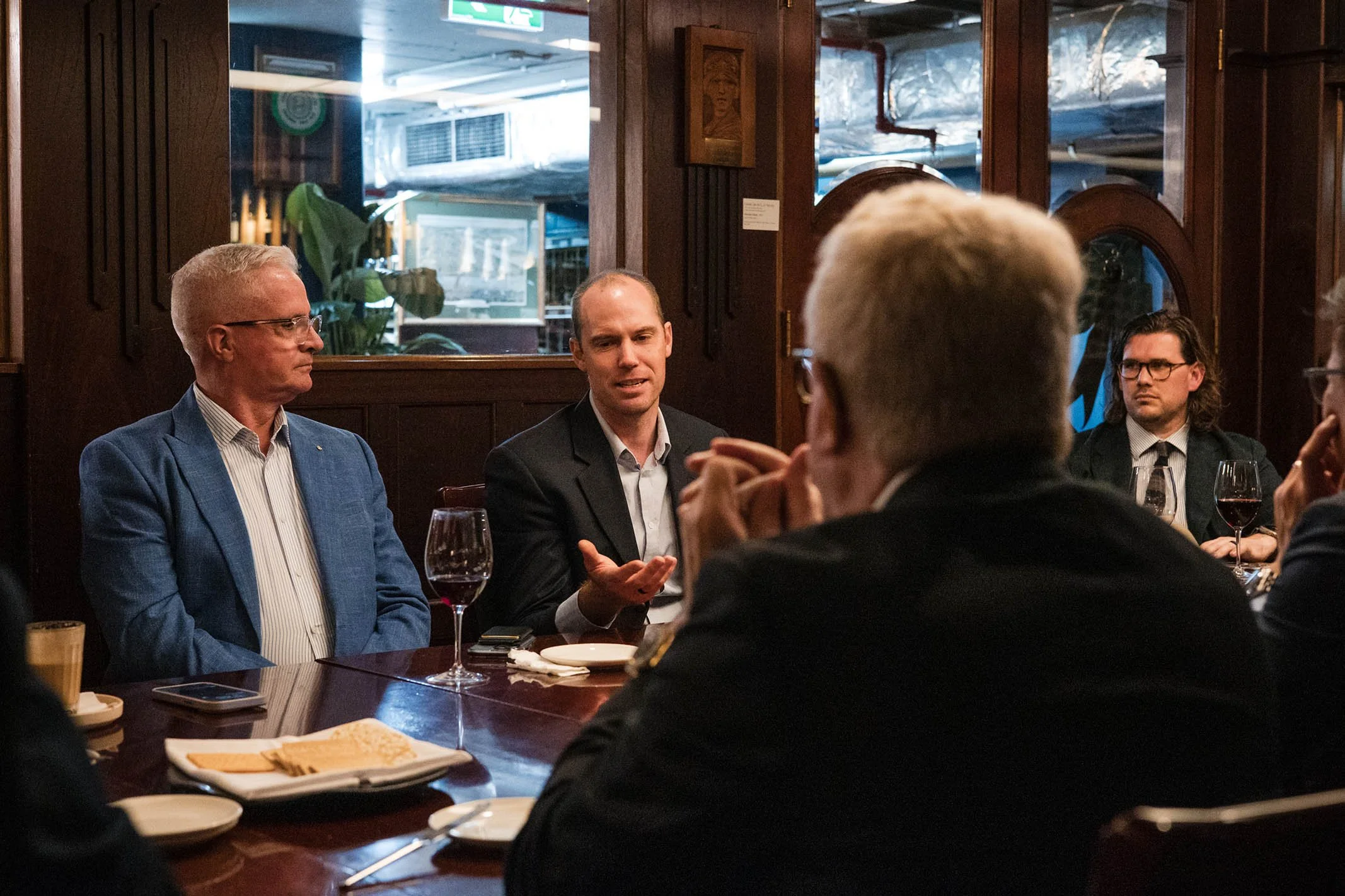 Group of people having a discussion in a restaurant, with one person speaking and others listening, with wine glasses and plates on the table.
