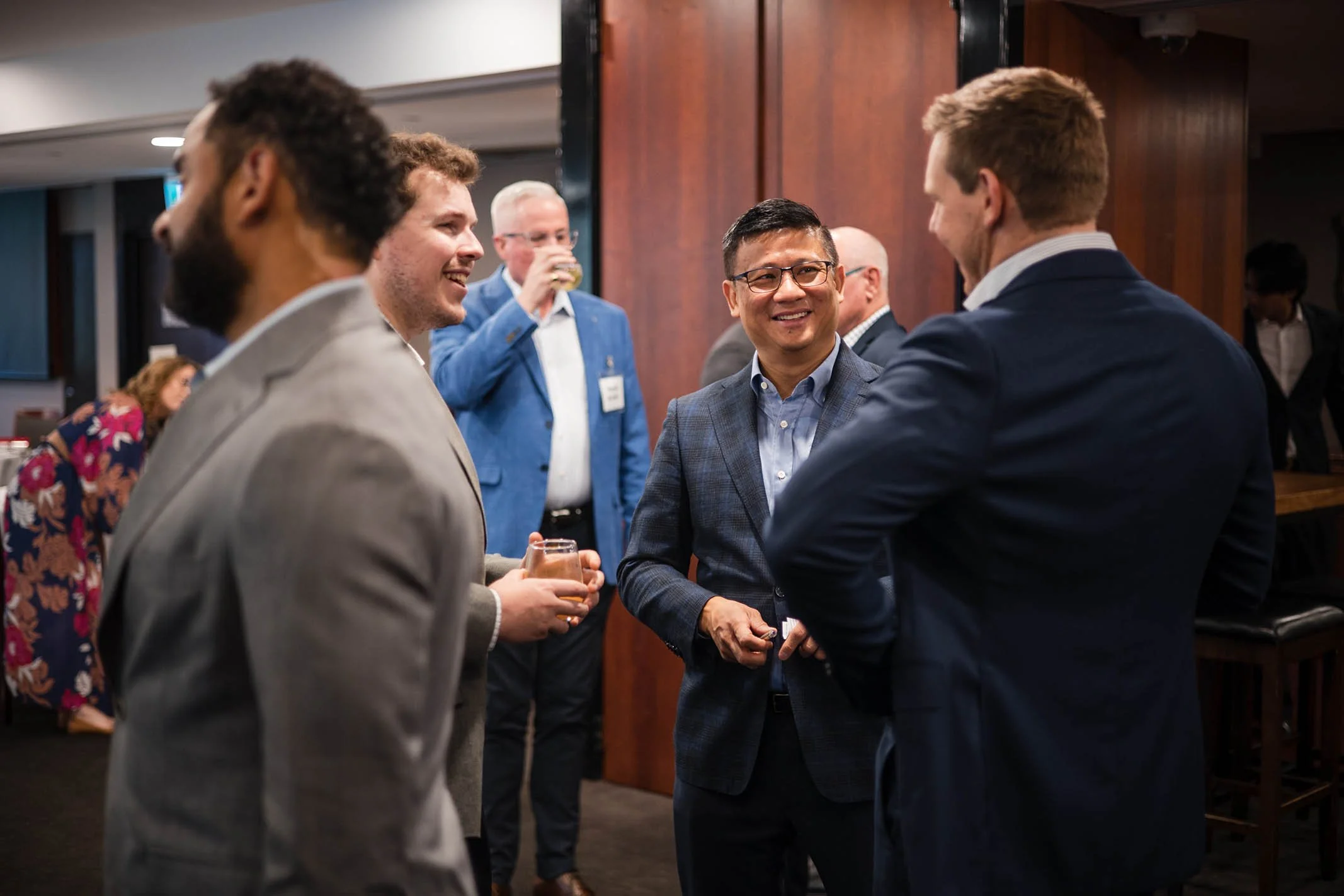 Group of professionally dressed men at a networking event, engaging in conversation and smiling, with a woman in floral dress in the background.