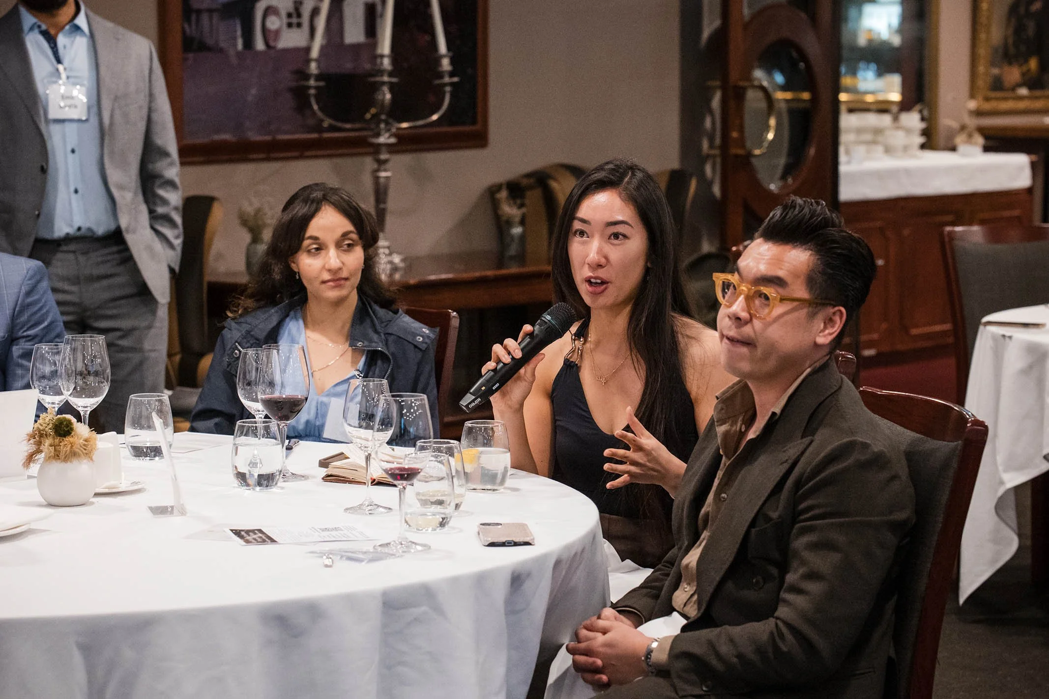 A woman is speaking into a microphone at a round table during a formal event, with others listening attentively. The table has wine glasses, a small vase with dried flowers, and a smartphone.