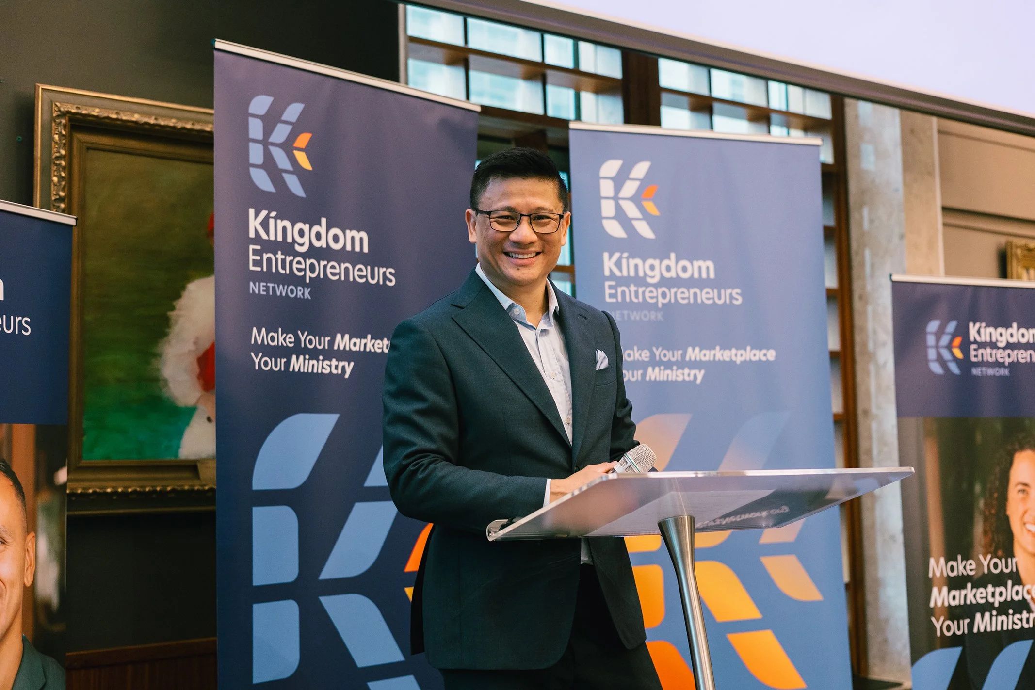 A man in a suit standing behind a podium, smiling, at a Kingdom Entrepreneurs Network event with banners in the background.