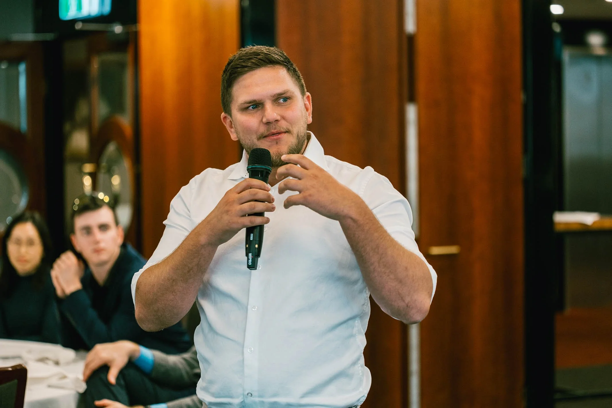 Man in white shirt holding a microphone and speaking at an indoor event, with seated audience members in the background.