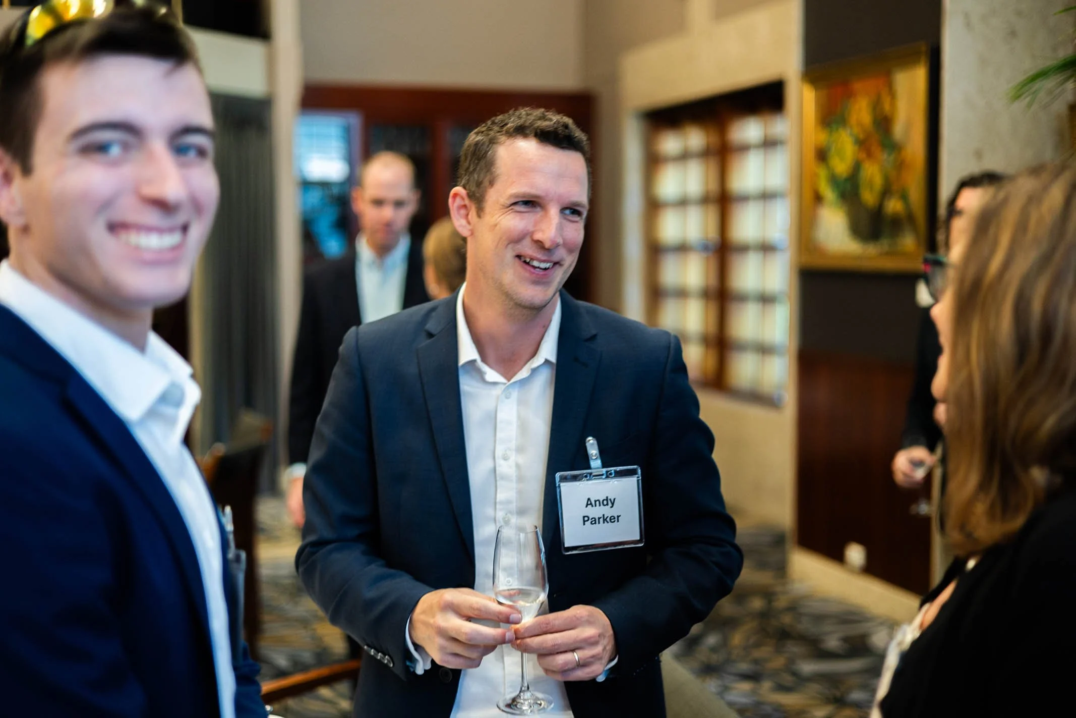 Man with a name tag labeled 'Andy Parker' holding a glass of champagne chatting with two women at a social event.