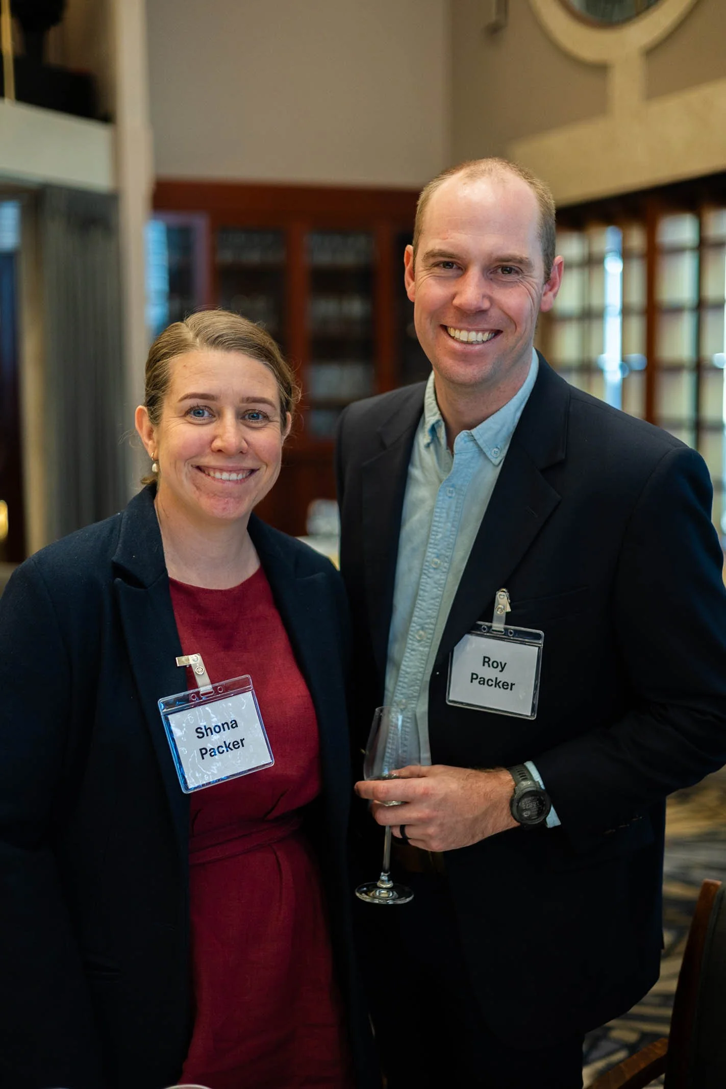 A man and woman smiling at a social event, both wearing name tags, with the woman’s tag reading 'Shona Packer' and the man’s 'Roy Packer.' The man is holding a glass of white wine.