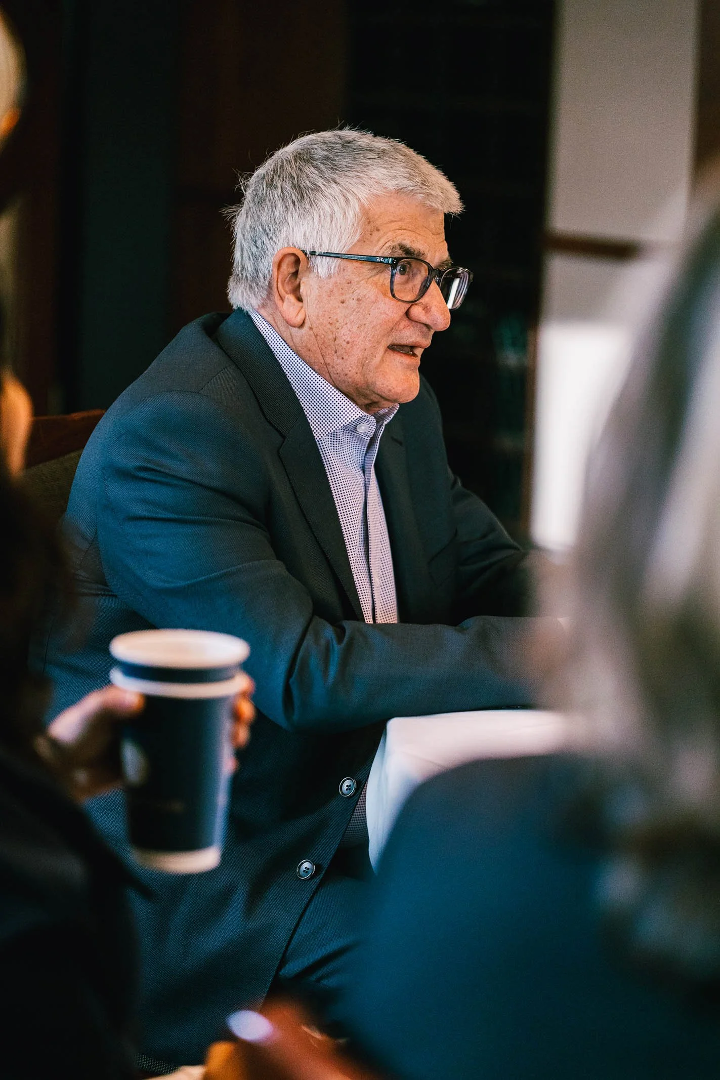 An older man with glasses and gray hair, wearing a dark suit and a light shirt, sitting at a table during a meeting or conference, with people holding coffee cups nearby.