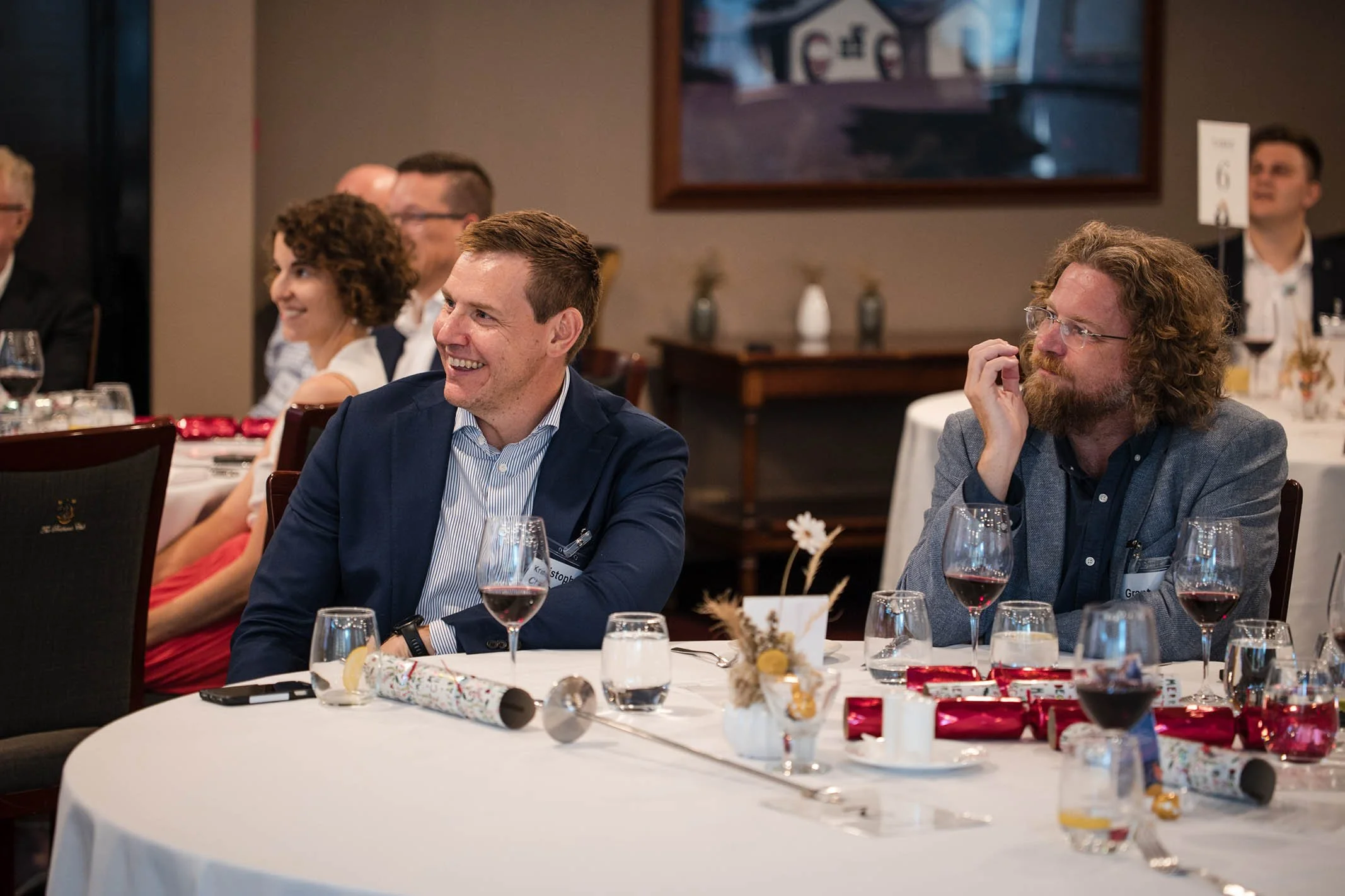 People sitting at a banquet table during an event, smiling and listening, with wine glasses and holiday decorations on the table.