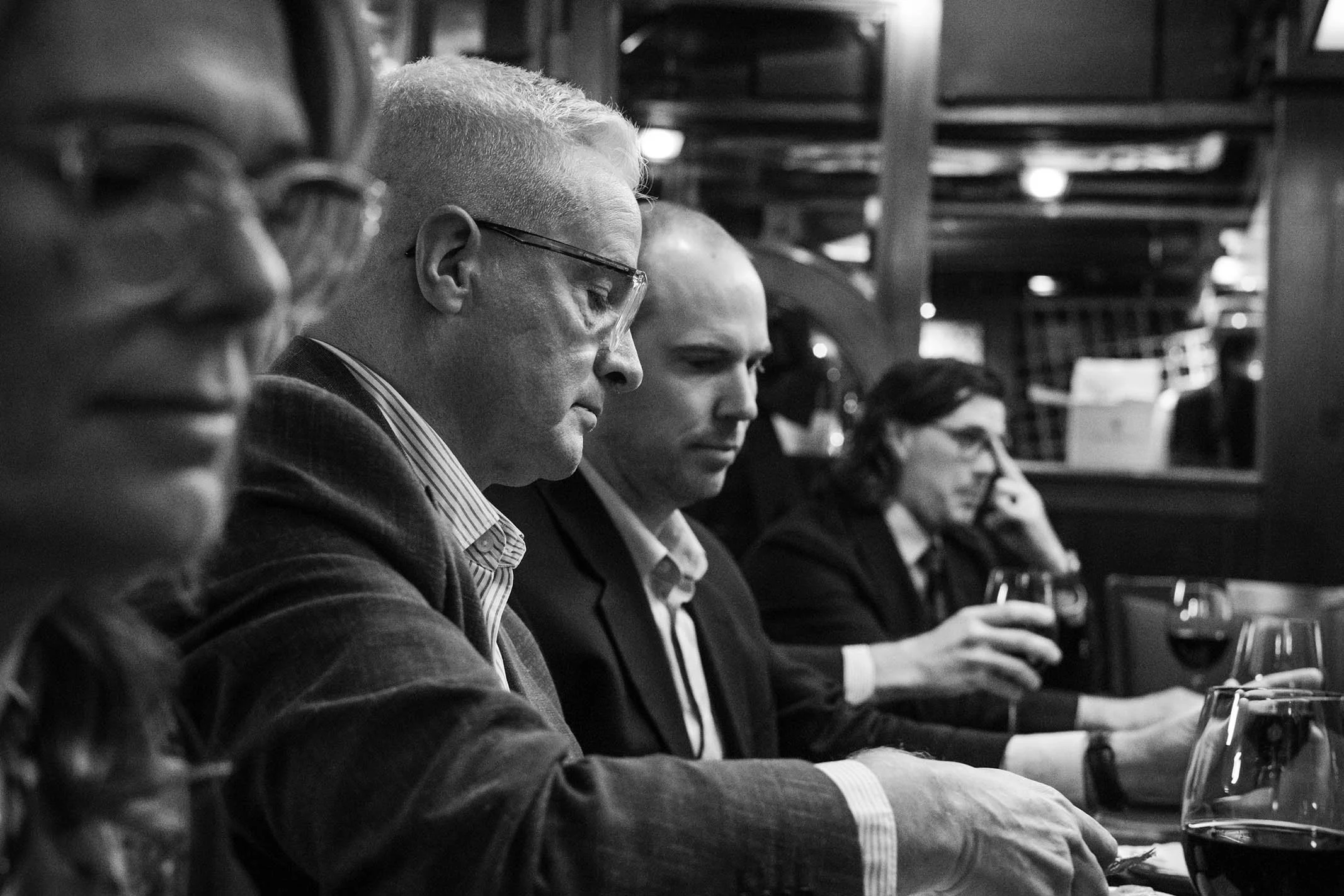 Four adults sitting at a table, three men and one woman, engaged in a serious or contemplative activity, with glasses of wine in front of them, in a dimly lit indoor setting.
