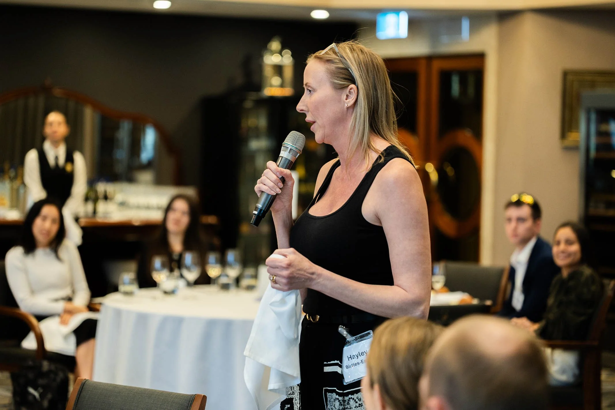 A woman speaking into a microphone at a formal event, with people seated at a round table listening.