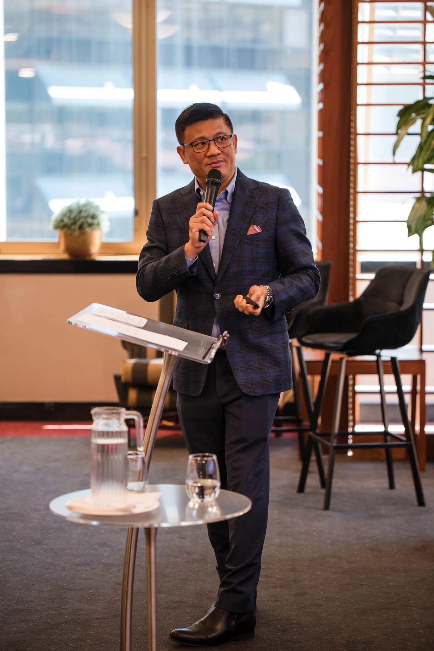 A man in a dark suit and glasses giving a presentation with a microphone in a room with large windows, a table with water pitcher and glasses, and chairs.