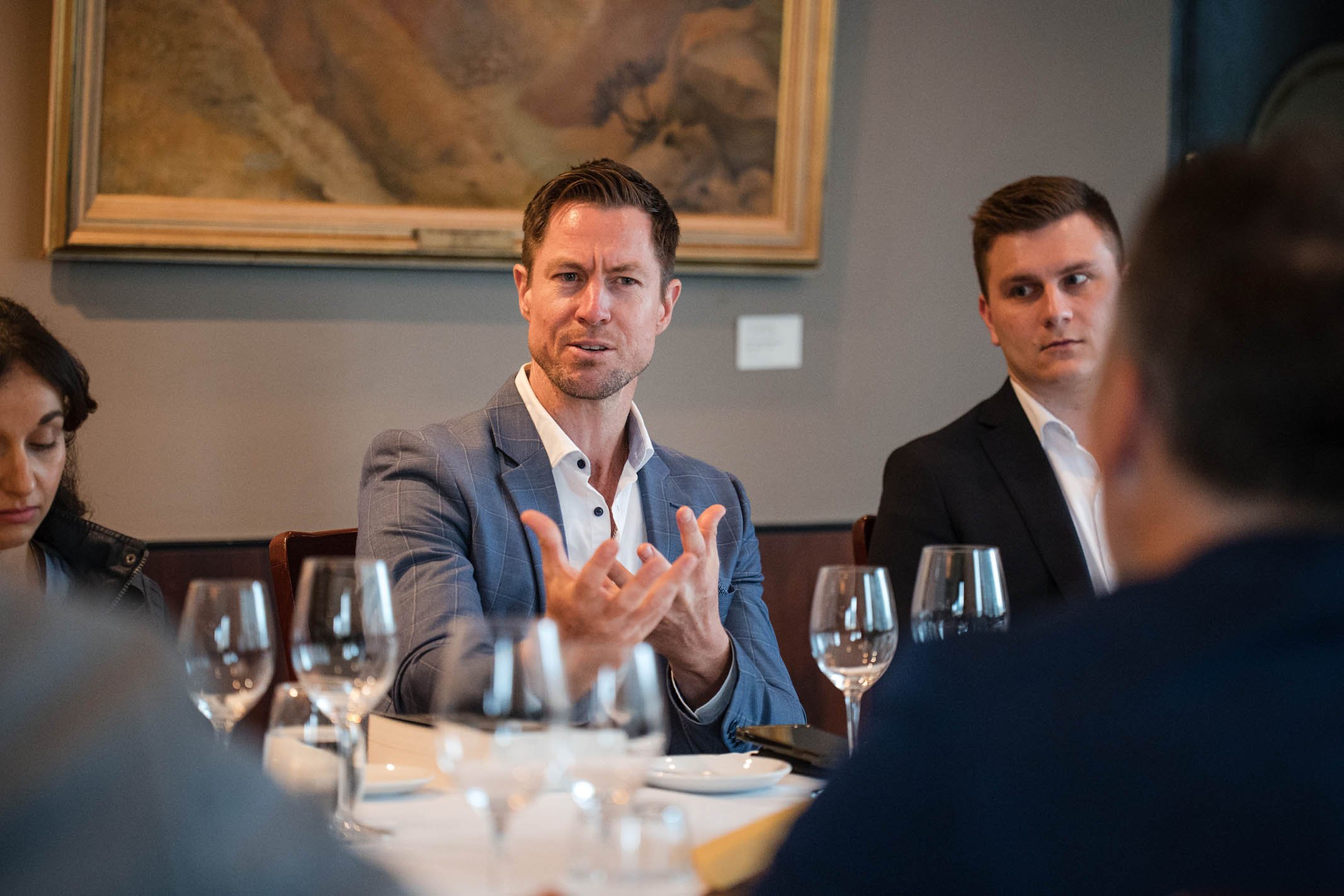 Man in a gray suit speaking during a meeting at a restaurant or conference room, with other people seated at the table, glasses and plates in front of him.