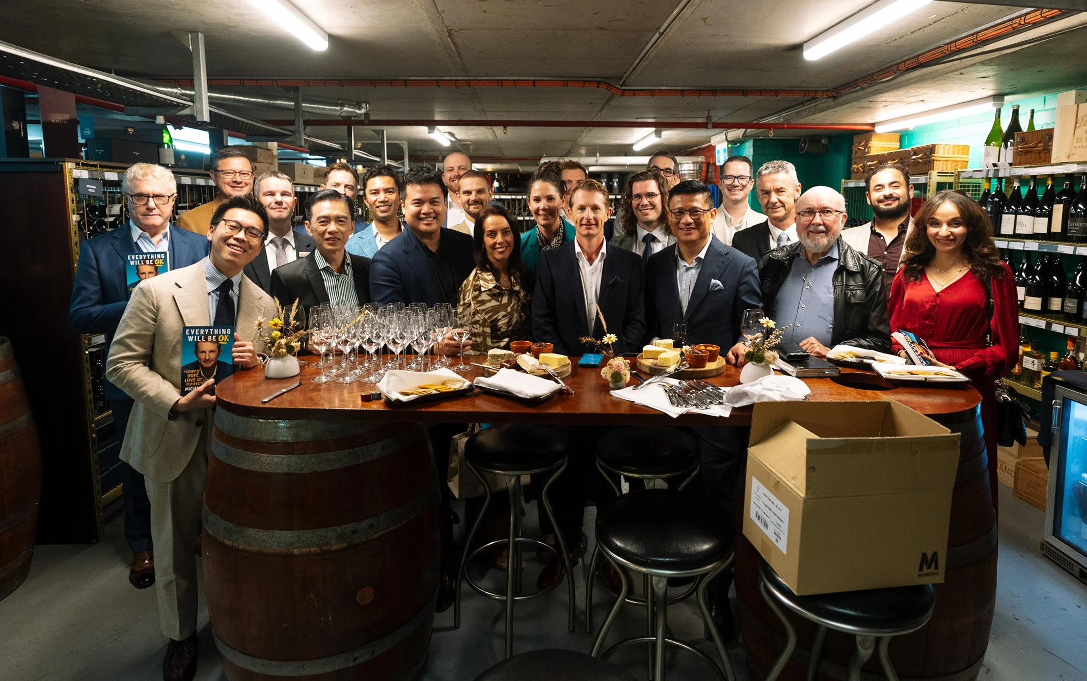 A group of people gathered around a wine barrel bar in a wine cellar or storage room, with wine bottles on shelves in the background. Some are holding books, and there are wine glasses and snacks on the table.