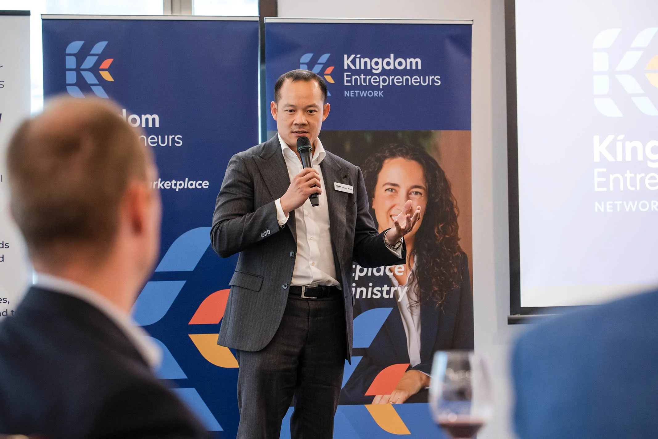 A man in a dark gray suit, white shirt, speaking into a microphone at a conference. Behind him are two banners with the logo and name of the Kingdom Entrepreneurs Network, one with a woman’s portrait and the other displaying the organization’s brandi