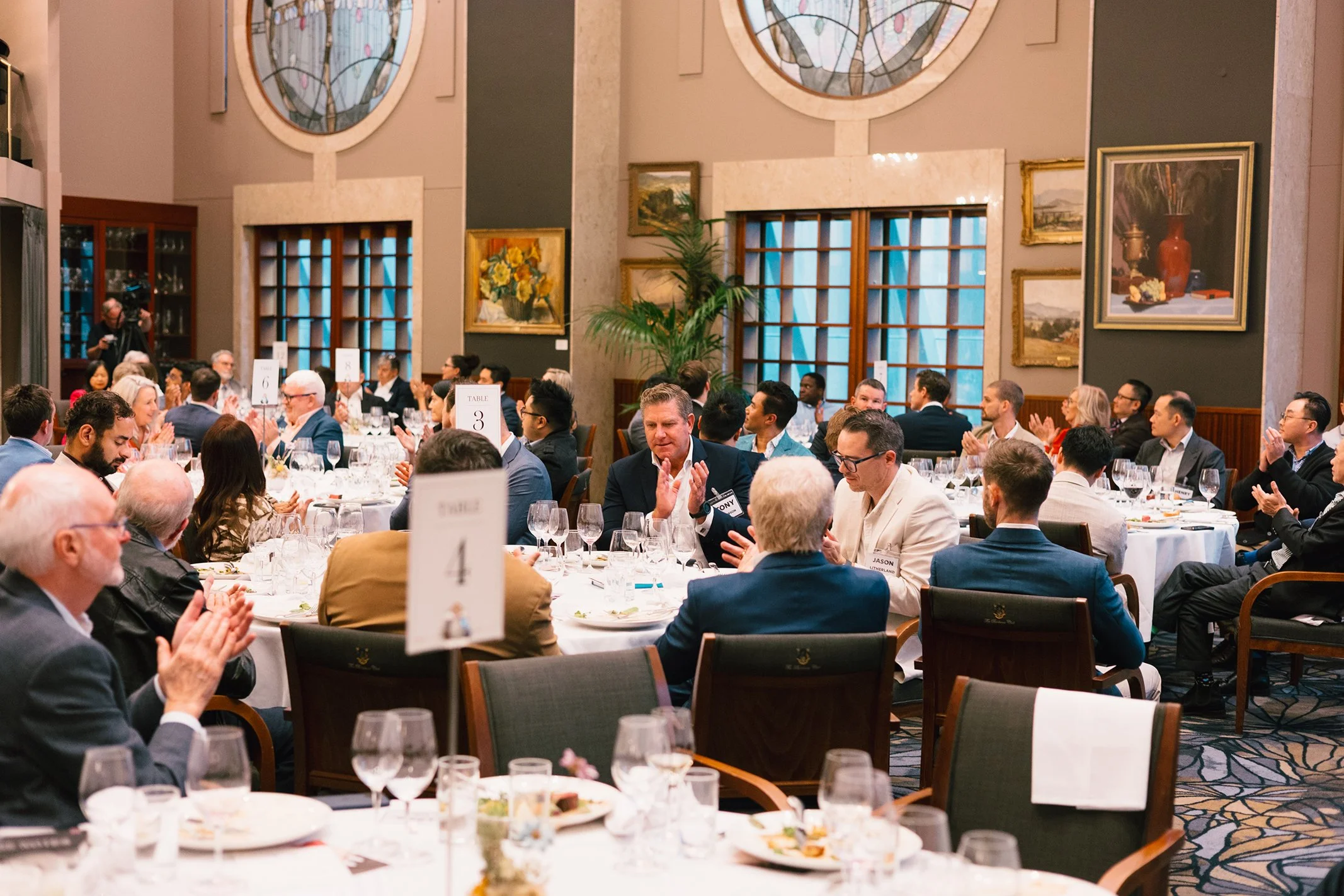 A formal dinner event with attendees sitting at round tables, applauding. The room features large stained glass windows, framed paintings, and decorative plants.