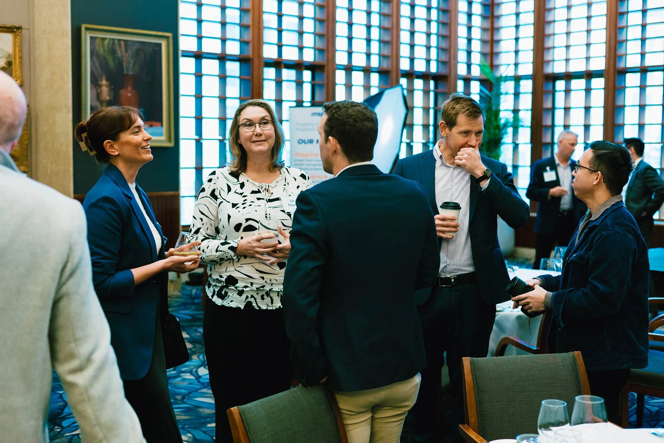 Group of six people engaging in conversation at a professional event or conference in a room with large windows and artwork on the walls.