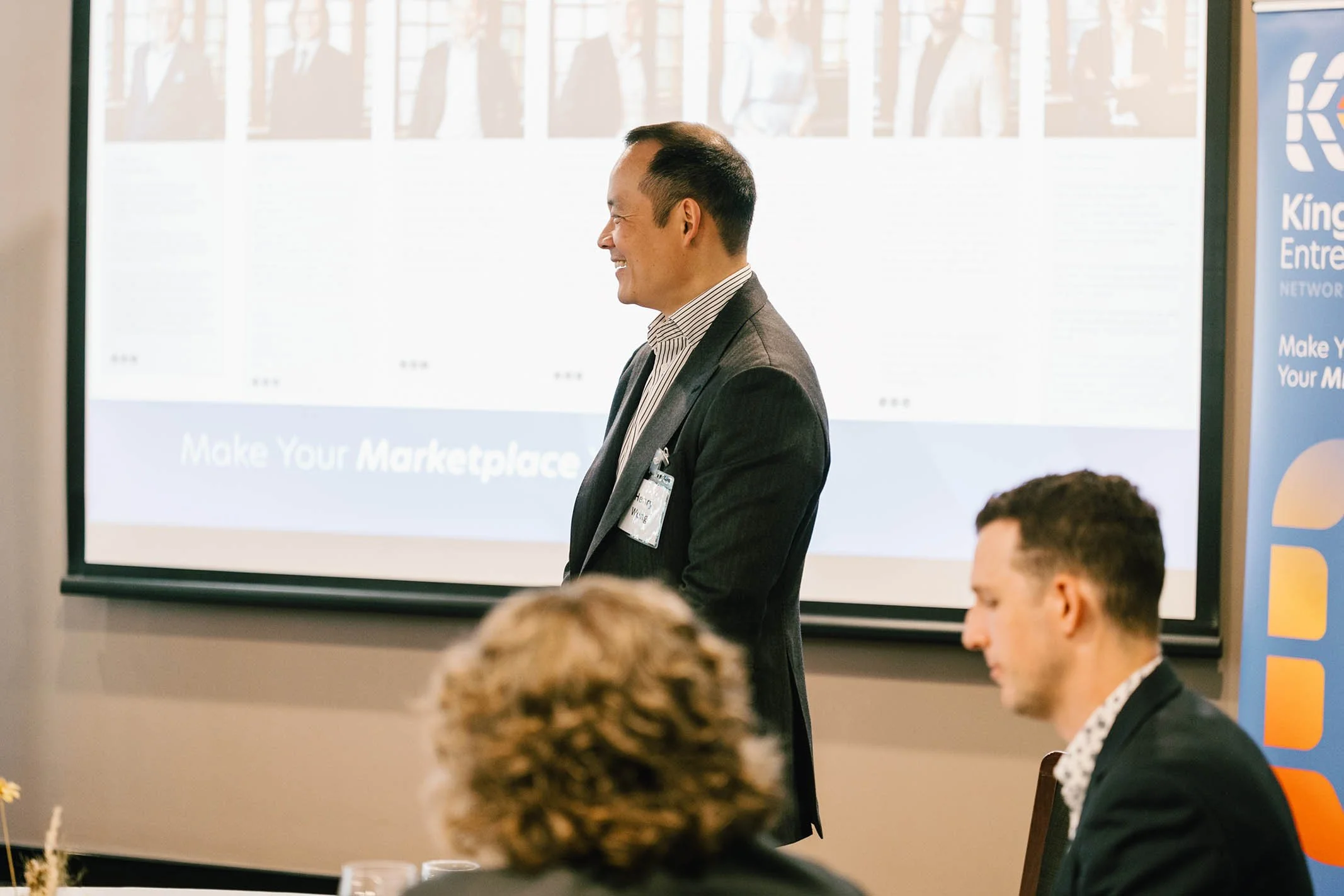 A man in a suit standing in front of a presentation screen, smiling during a conference or seminar.
