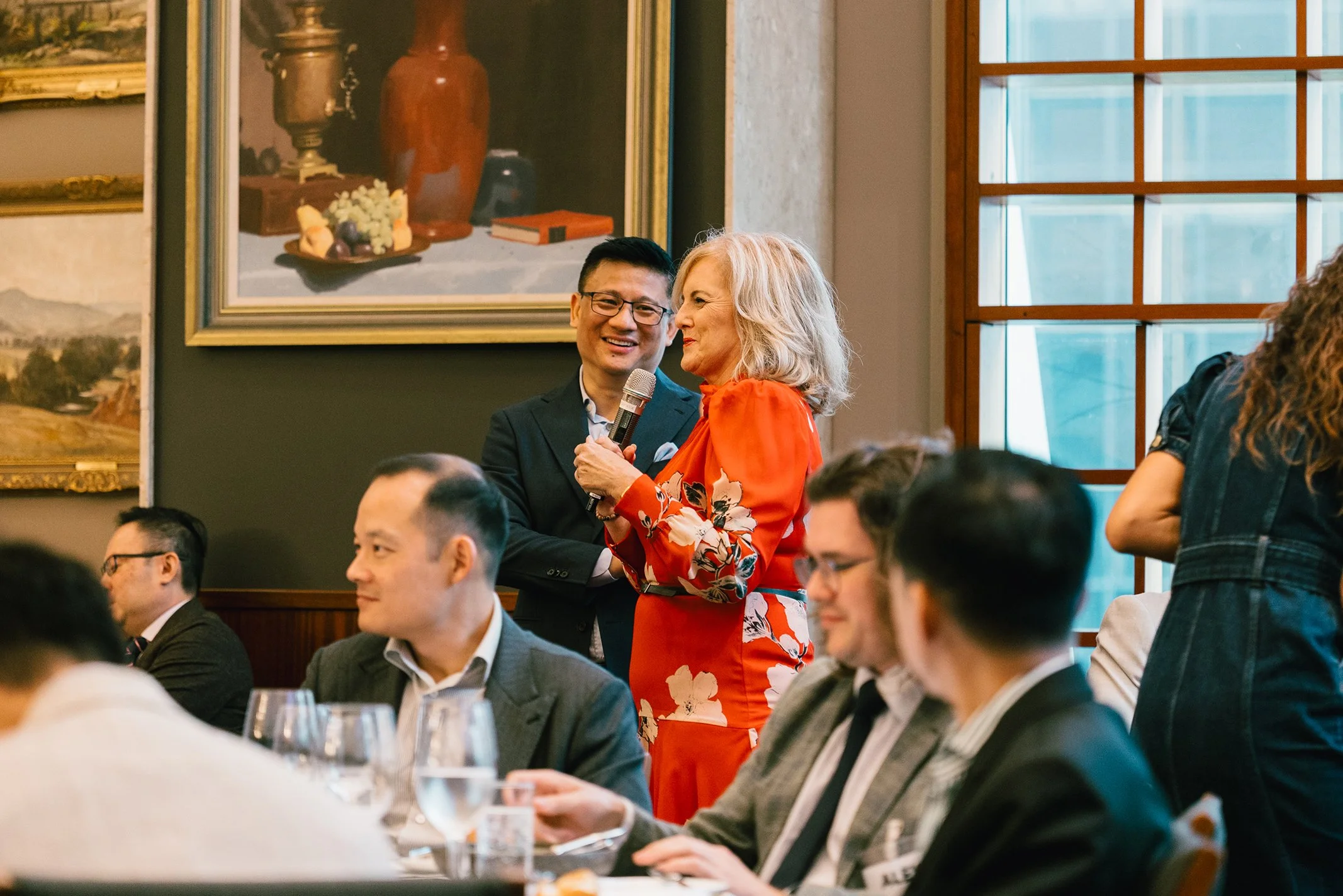 A woman in a red floral dress holding a microphone and smiling as she chats with a man in a dark suit at a formal event. Several people seated at a table in front are visible, with glasses of water and dinnerware. The background features large painti