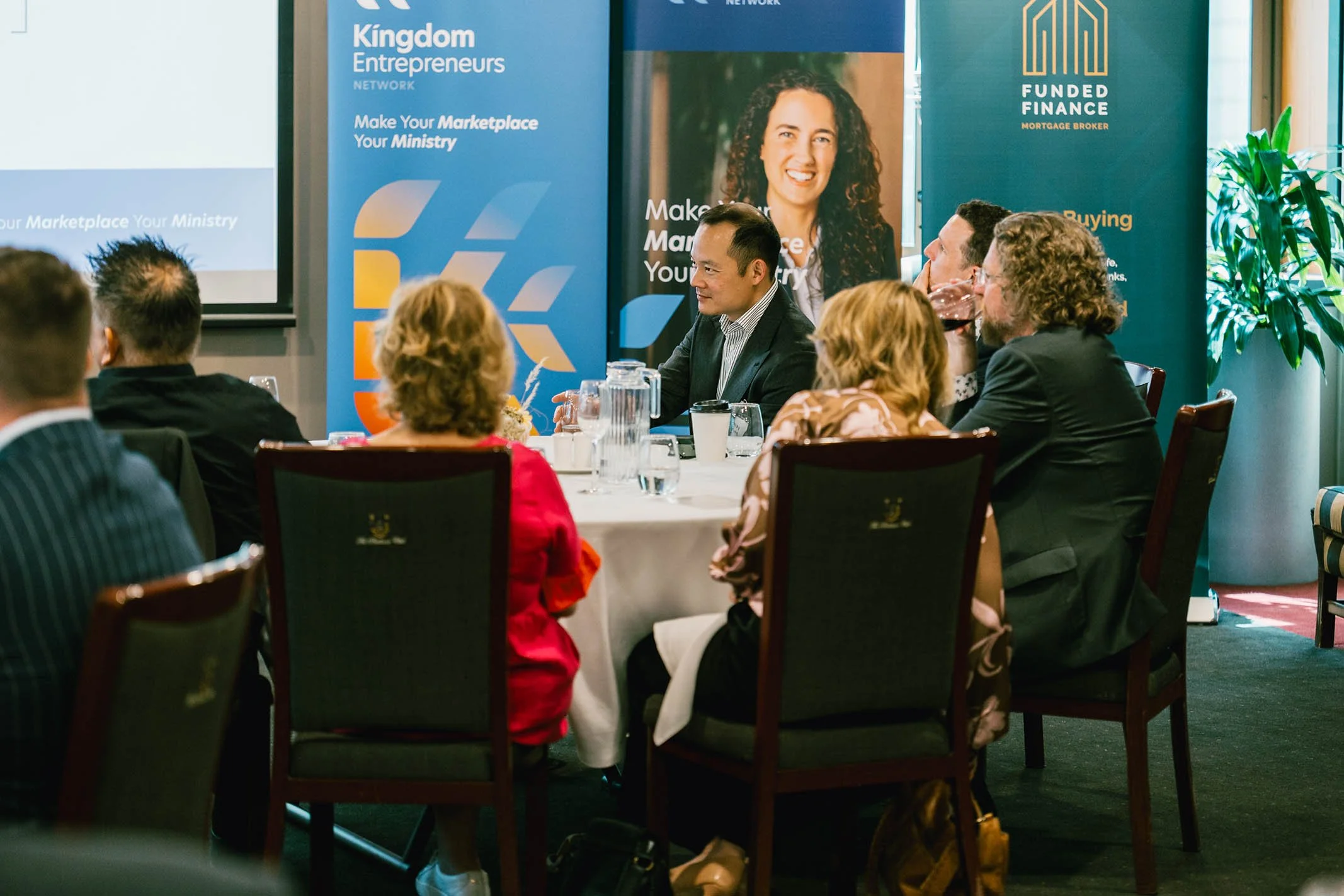 A group of business professionals attending a conference or meeting, seated around a round table with a presentation screen and banners in the background.